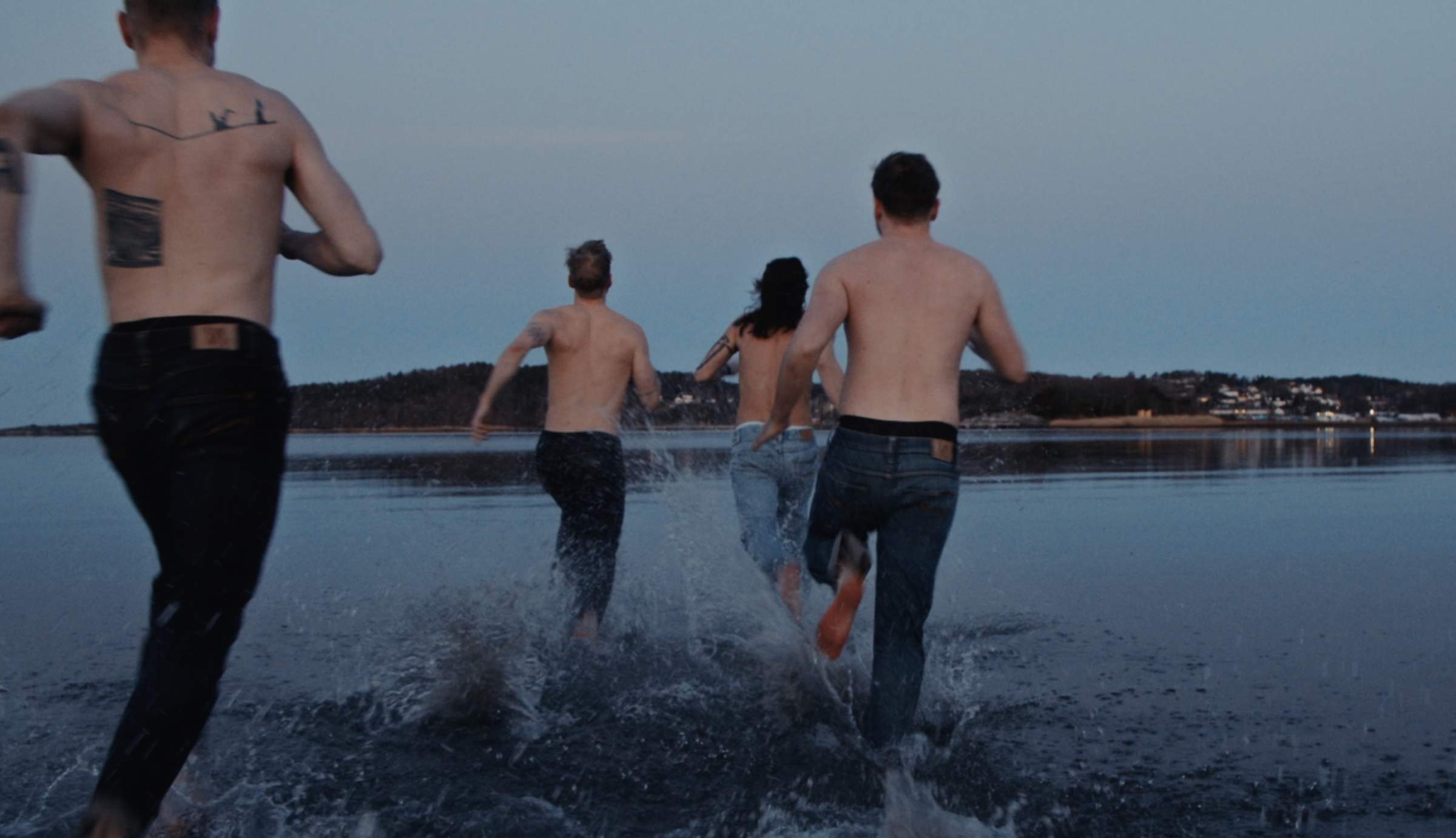 Four shirtless people run into a body of water at dusk, creating splashes as they move toward the distant shoreline.