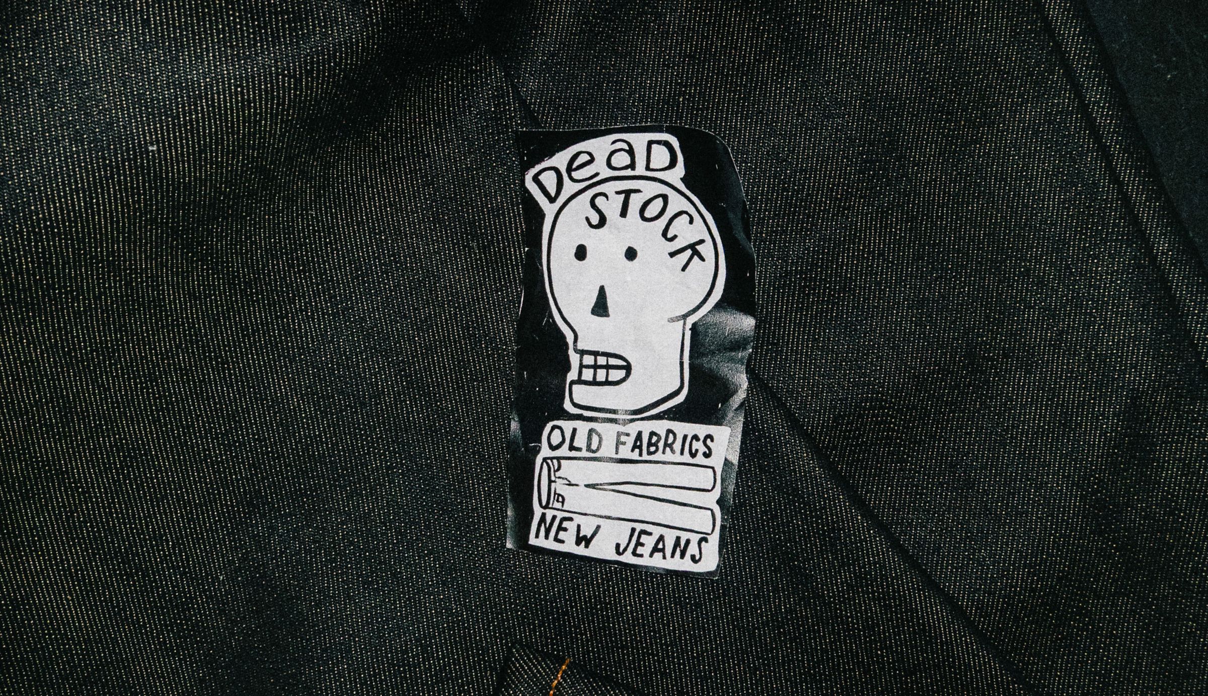 Sticker with a skull illustration and text: "Dead Stock, Old Fabrics, New Jeans" on a dark fabric background.