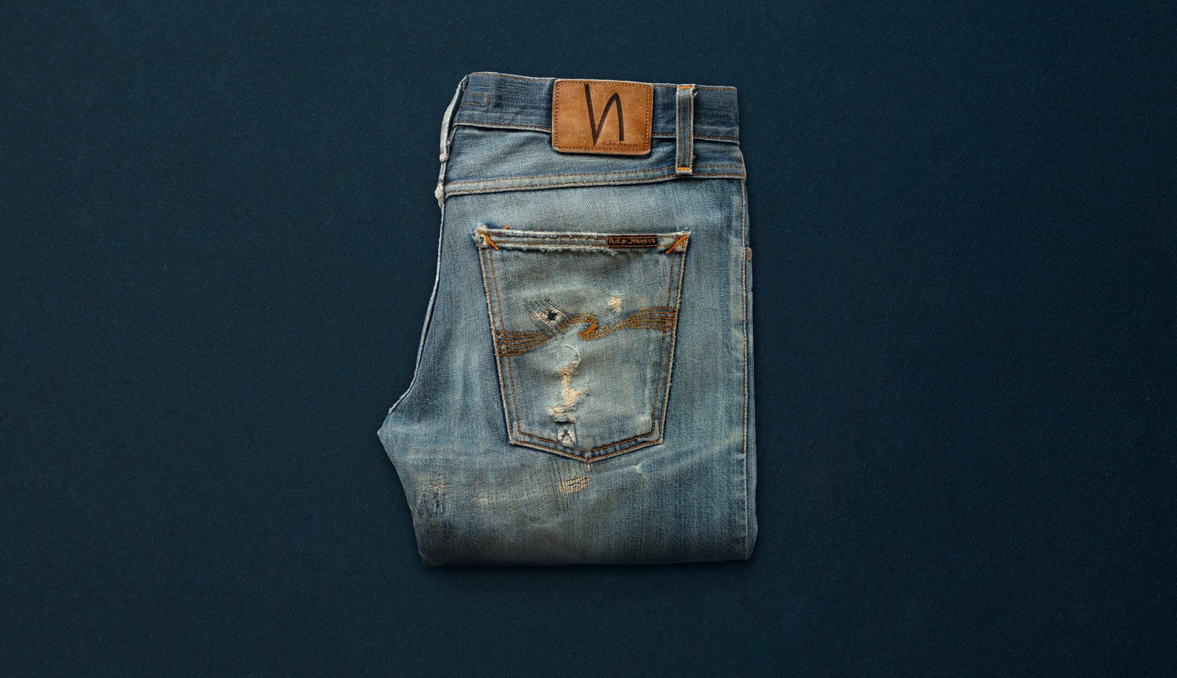 A pair of well used and repaired Nudie Jeans on a blue background..