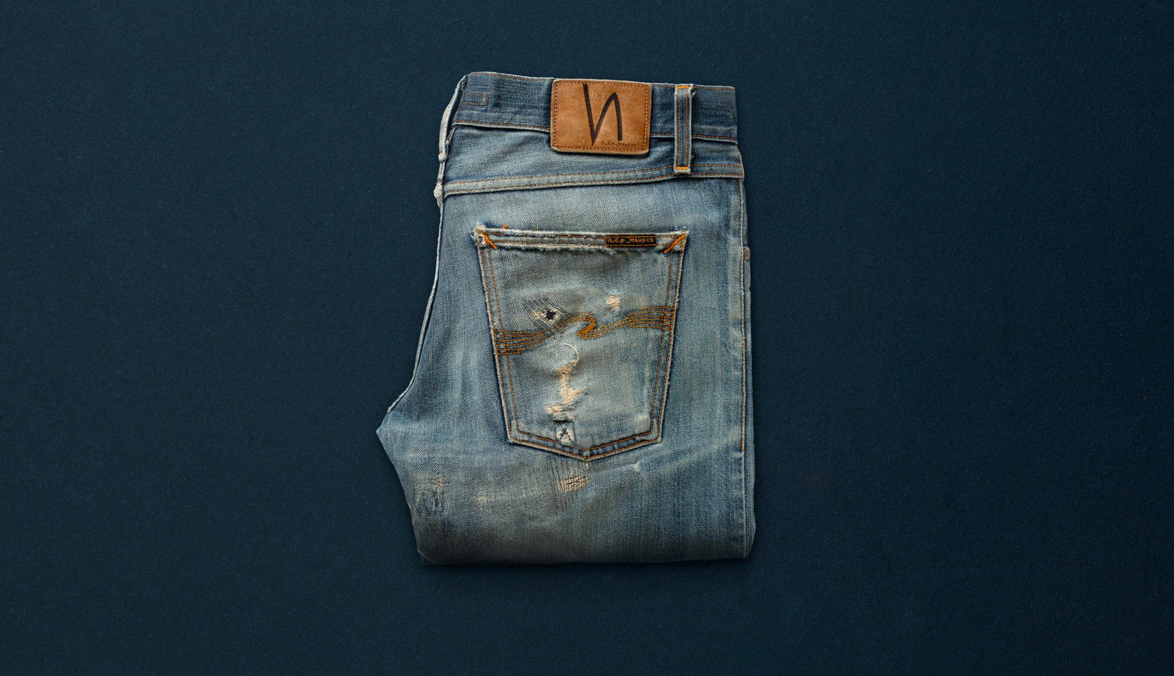 A pair of well used and repaired Nudie Jeans on a blue background..