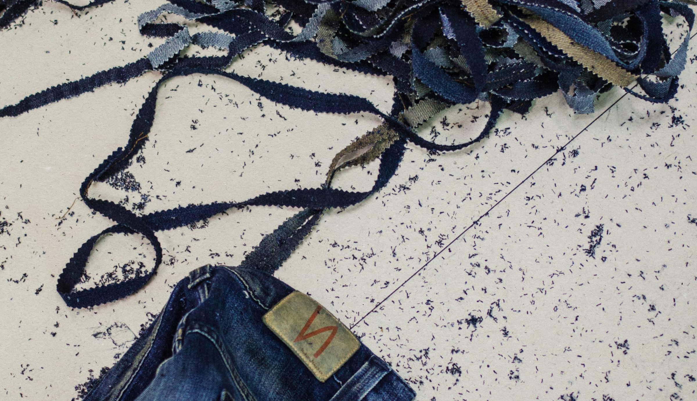Denim strips scattered on the floor, blue jeans showing a Nudie Jeans leather patch and stitching.