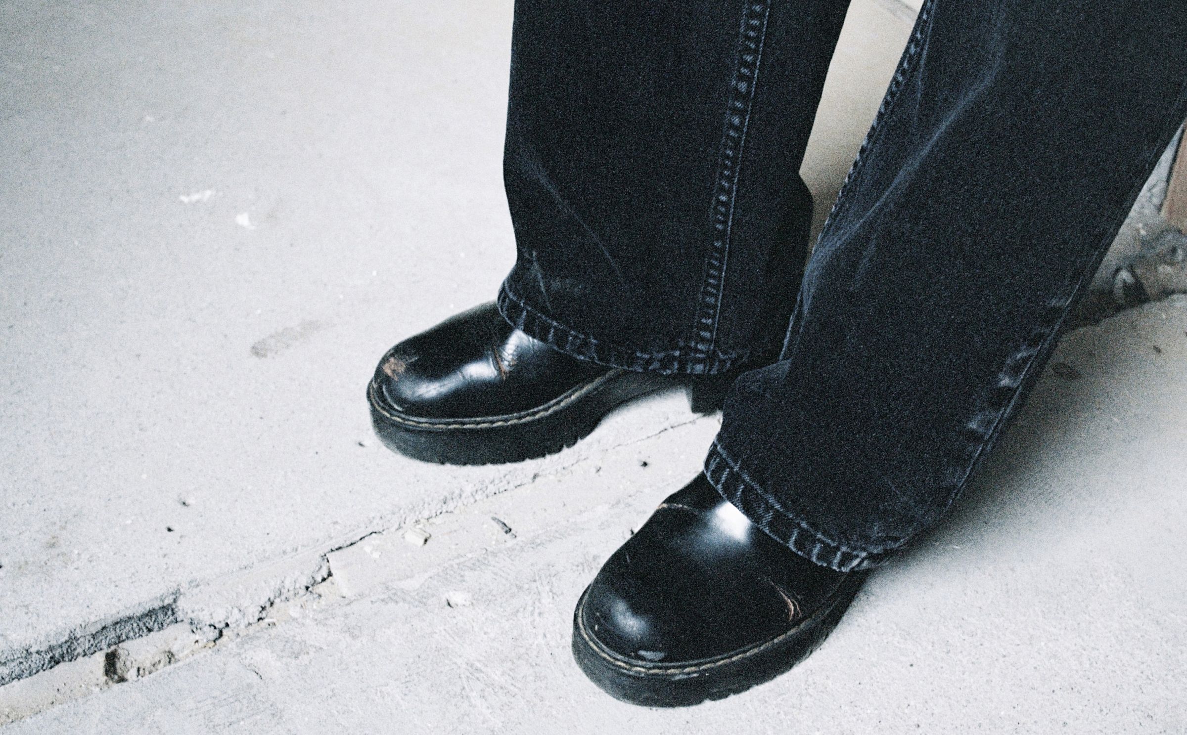 Person wearing black leather shoes and black denim jeans standing on a concrete floor.