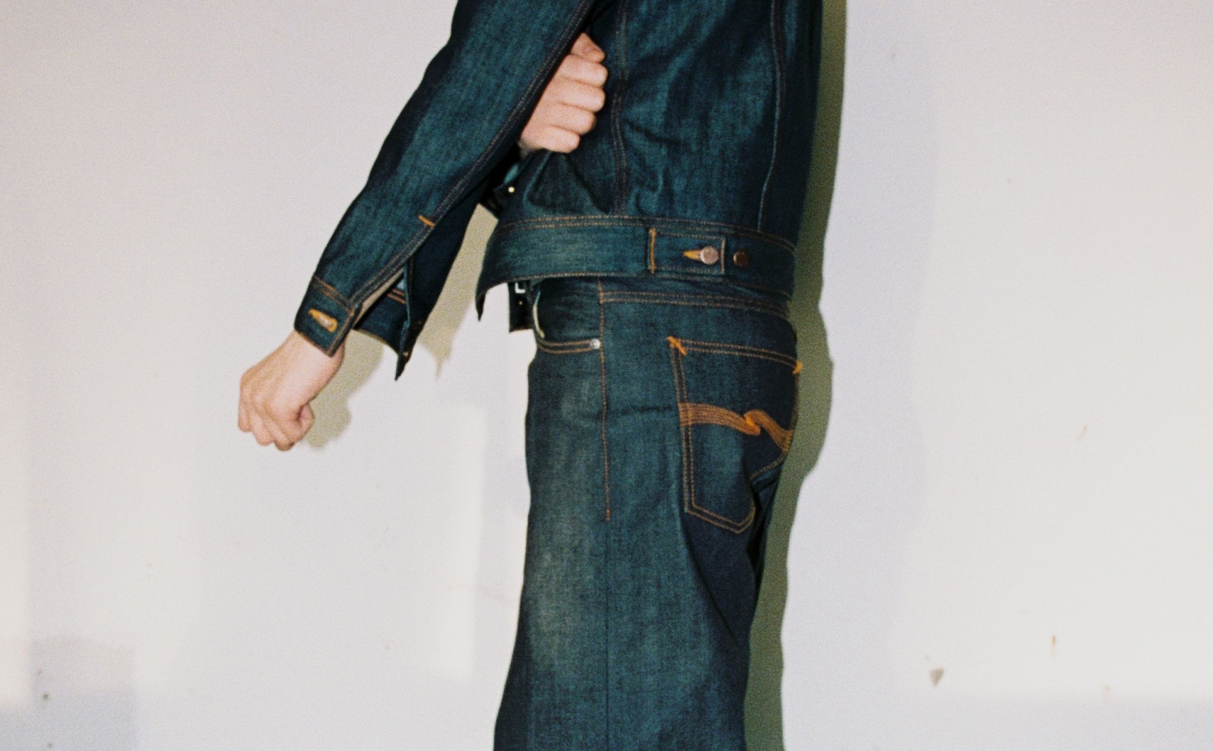 Person wearing a dry raw denim jacket and wide-leg jeans, standing against a plain wall with a yellow baseboard.