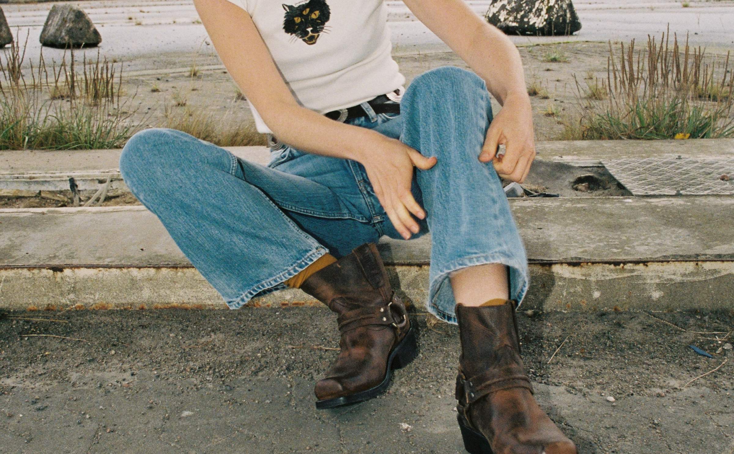 Model in light wash Nudie Jeans with white cat-print tee and brown harness boots, sitting on a curb.