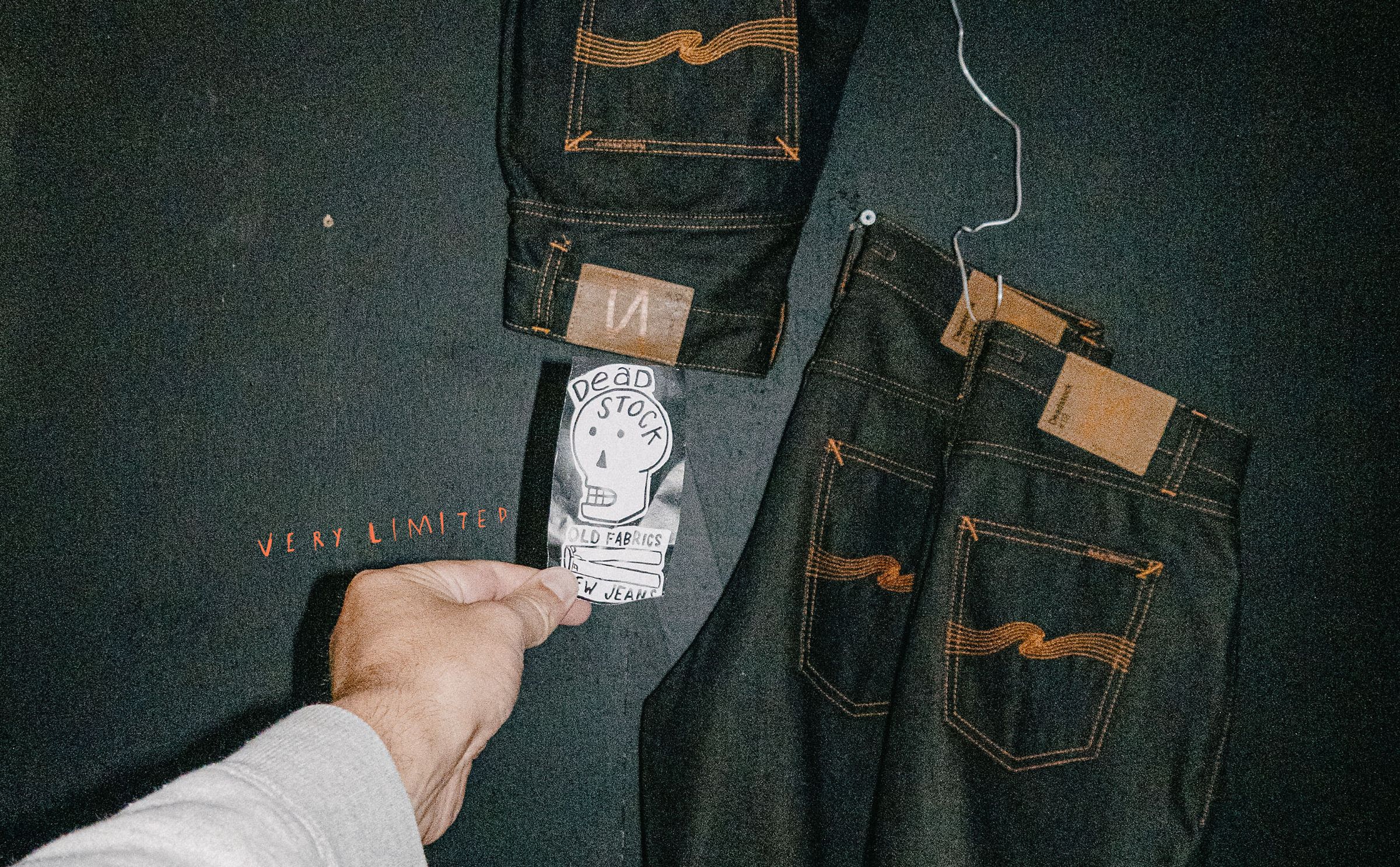 A hand holds a "Deadstock" tag in front of two pairs of dark denim jeans hanging on a black wall, with "Very Limited" written in red.