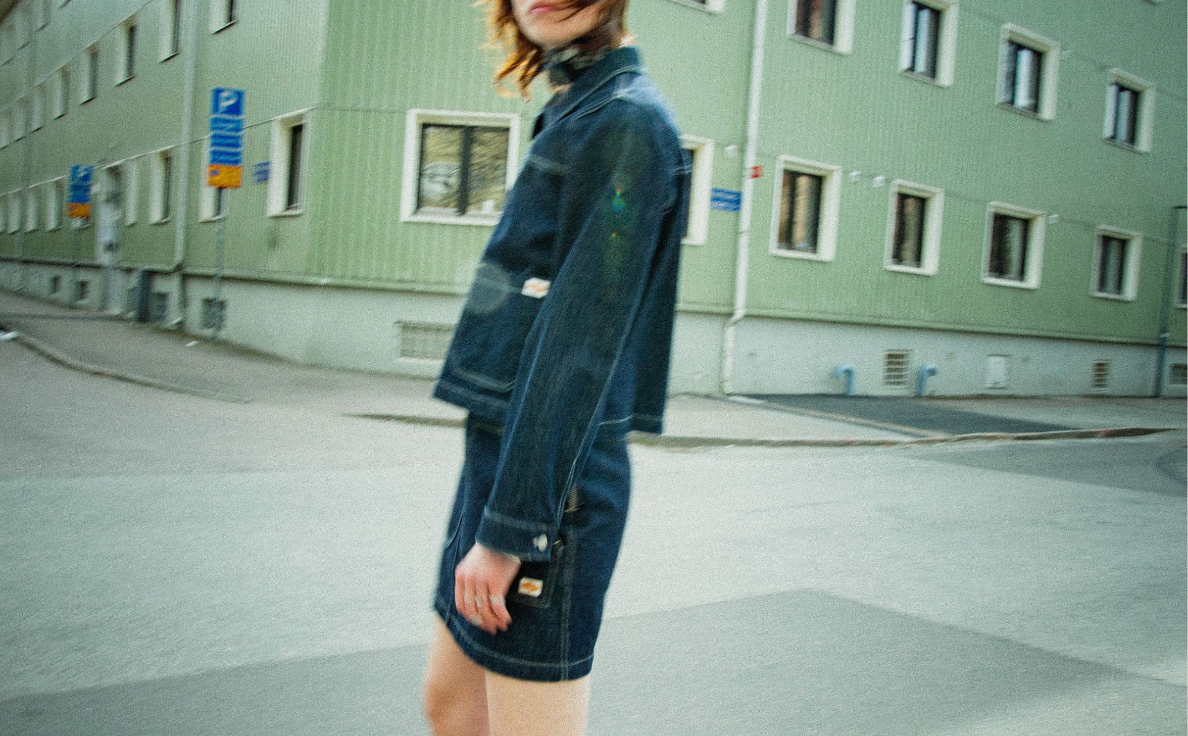 Person in a denim jacket and denim skirt walking on a street near a green building with multiple windows. Blurred motion effect.