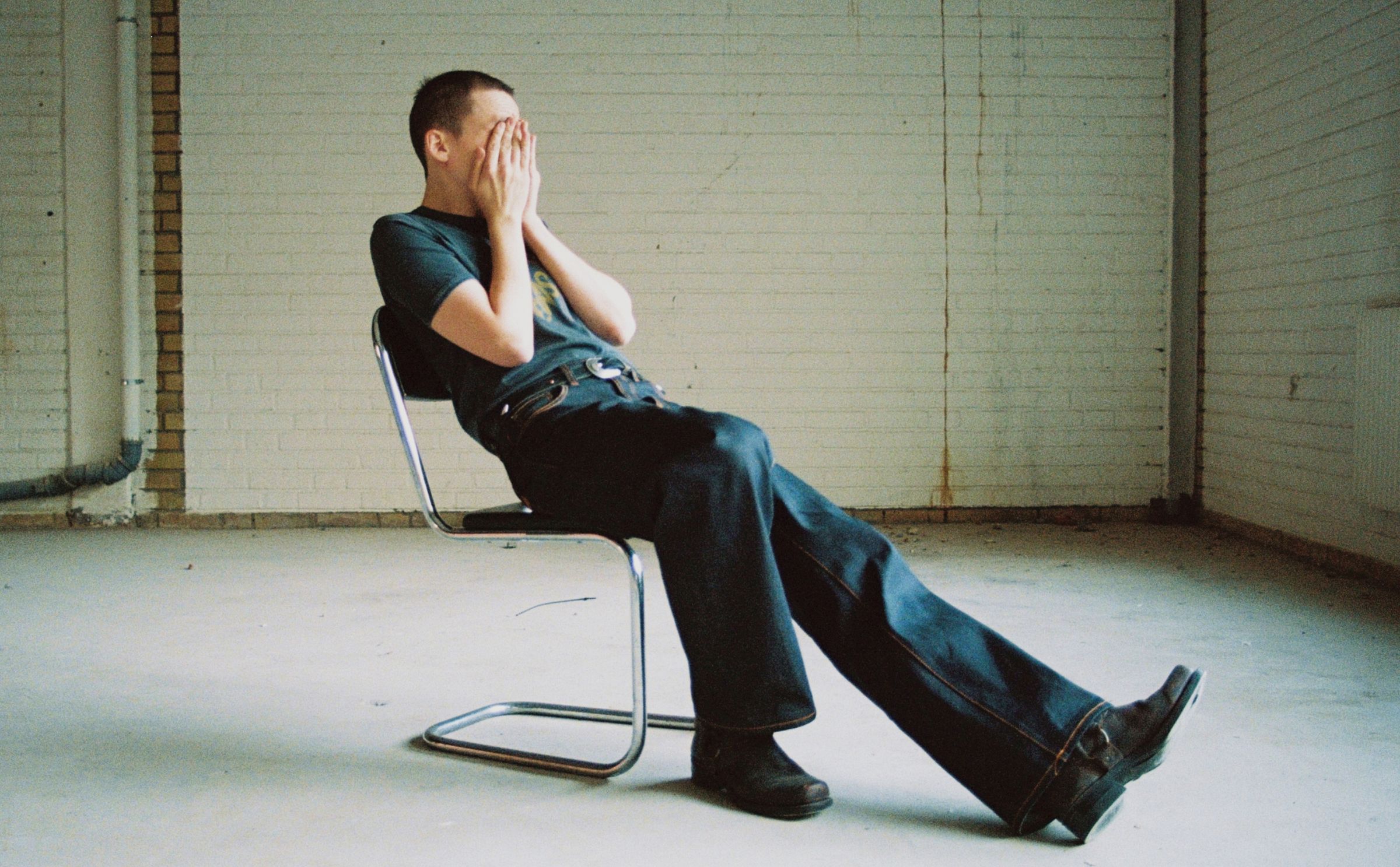 Person sitting on a chair, wearing dry raw jeans and shoes, covering their face with hands, against a white brick wall background.