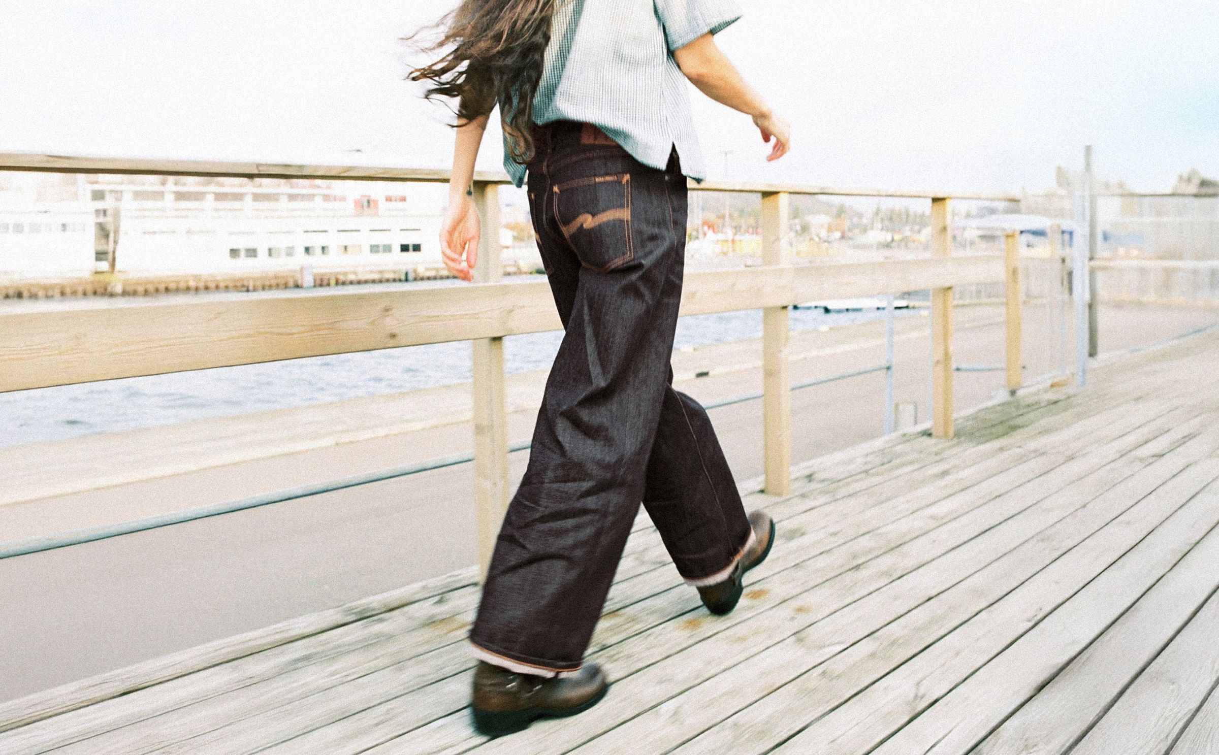 Person walking on a wooden boardwalk near water, wearing raw selvage jeans and boots, with long hair flowing, and a light shirt.