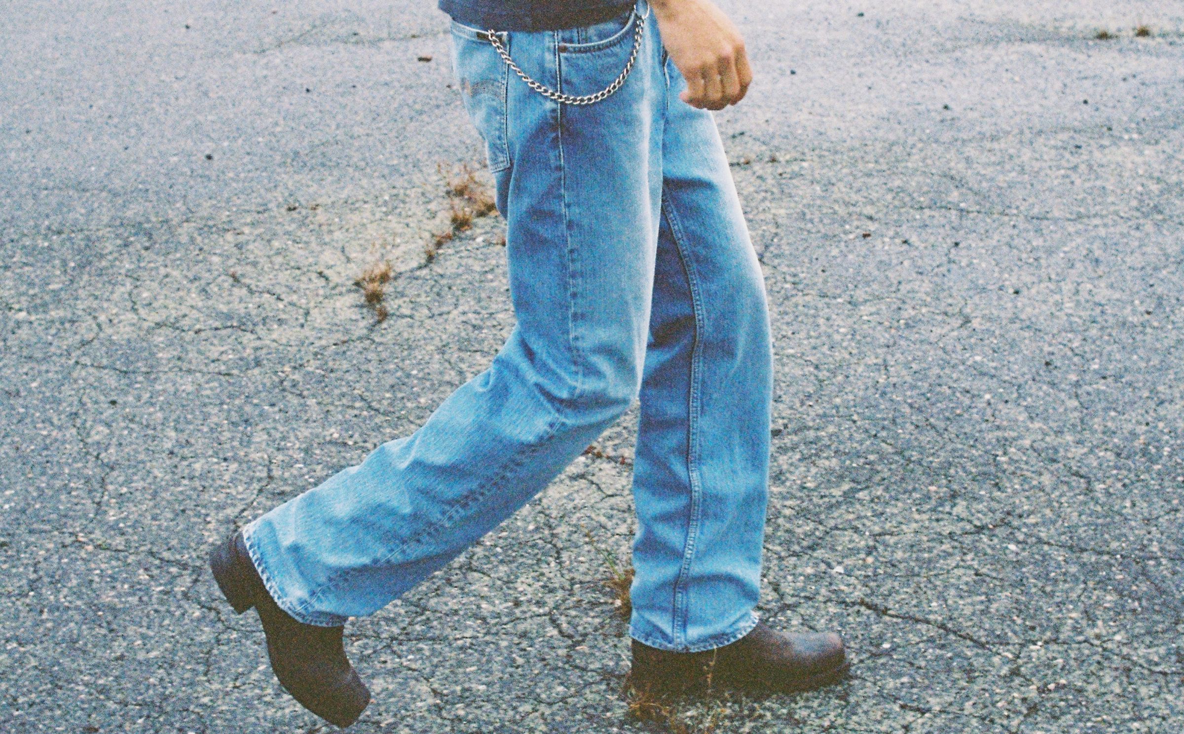 Person walking on a cracked pavement wearing light washed jeans with a chain and black boots.