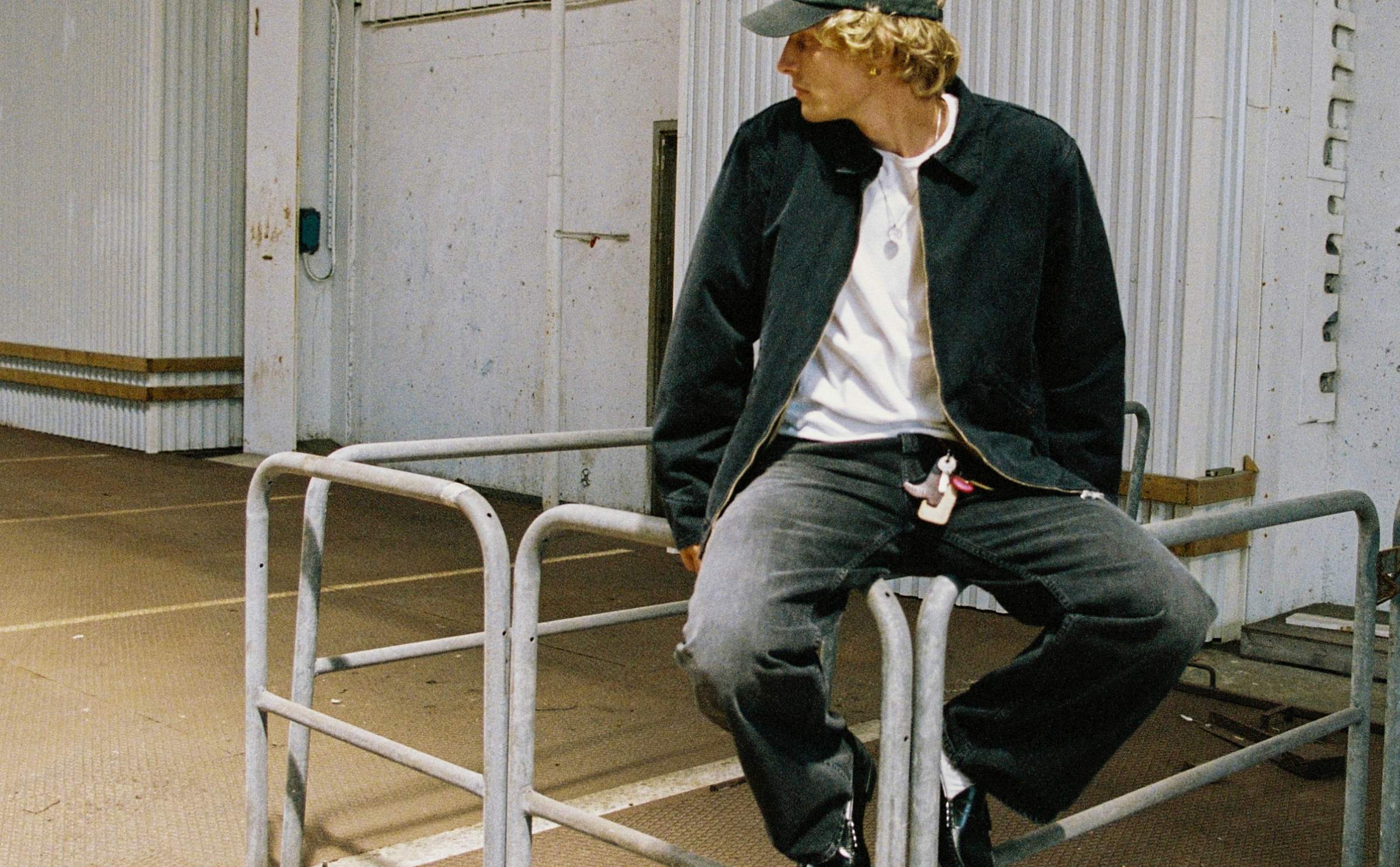 Model in black Nudie Jeans denim jacket and black jeans with white tee, sitting on metal railing outside warehouse.