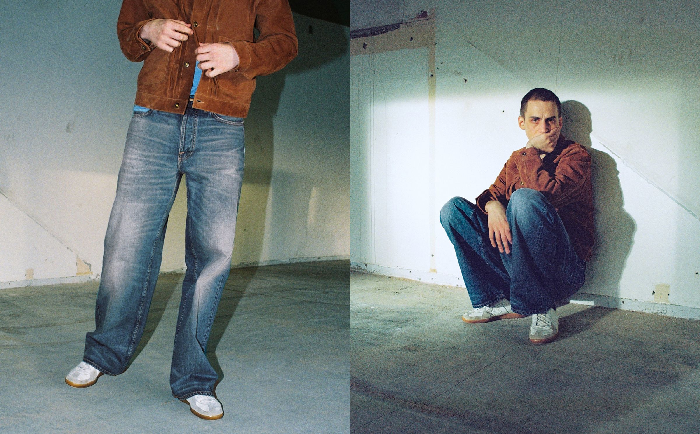 Man in a brown jacket and jeans stands and squats in a dimly lit, unfinished room with concrete floor and white walls.