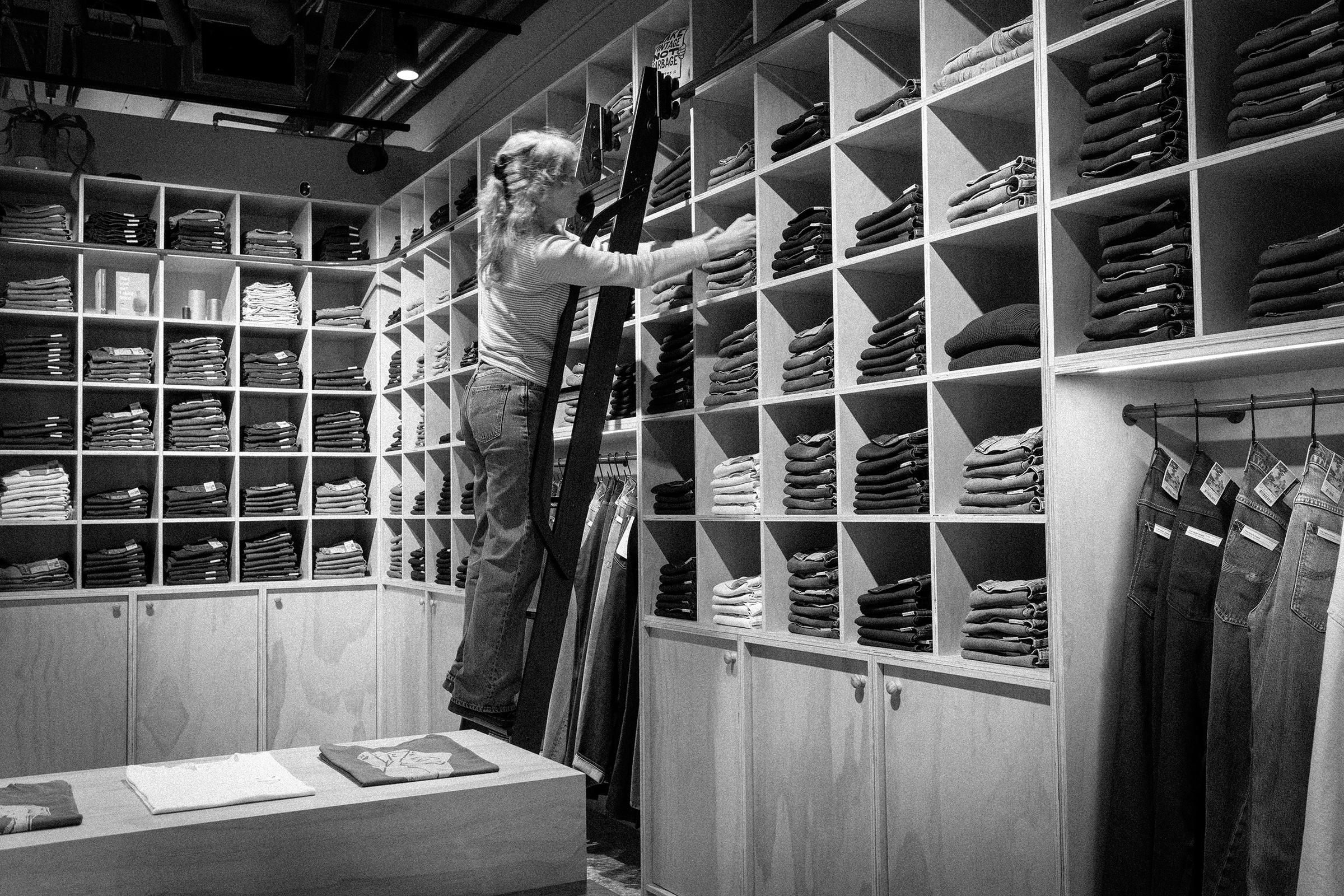 Person organizing folded clothes on shelves using a ladder in a retail store. The store is filled with neatly arranged garments.