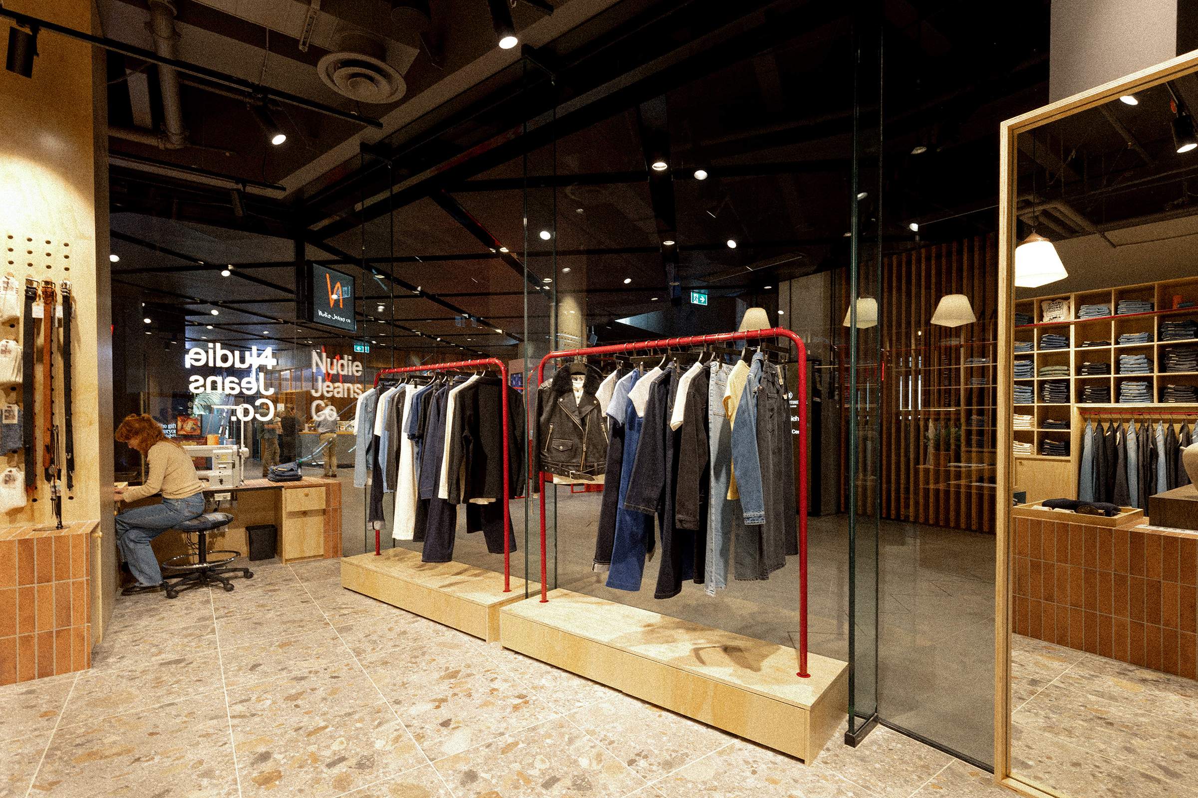 Clothing store interior with jeans and jackets on racks, a person working at a desk, and shelves of folded clothes in the background.