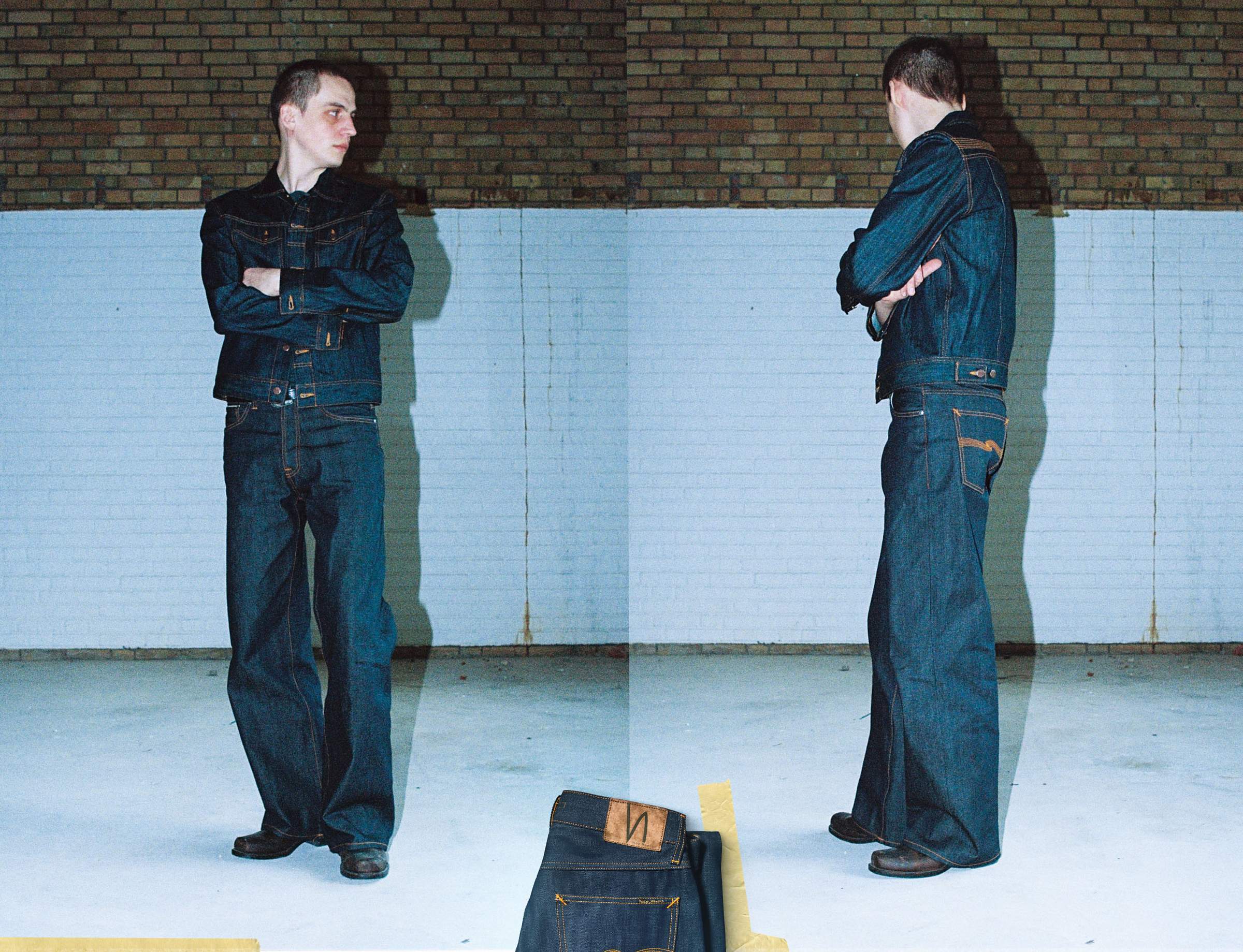 Person in dry, raw denim jacket and wide-leg jeans, standing with arms crossed, facing forward and sideways against a brick and white wall.