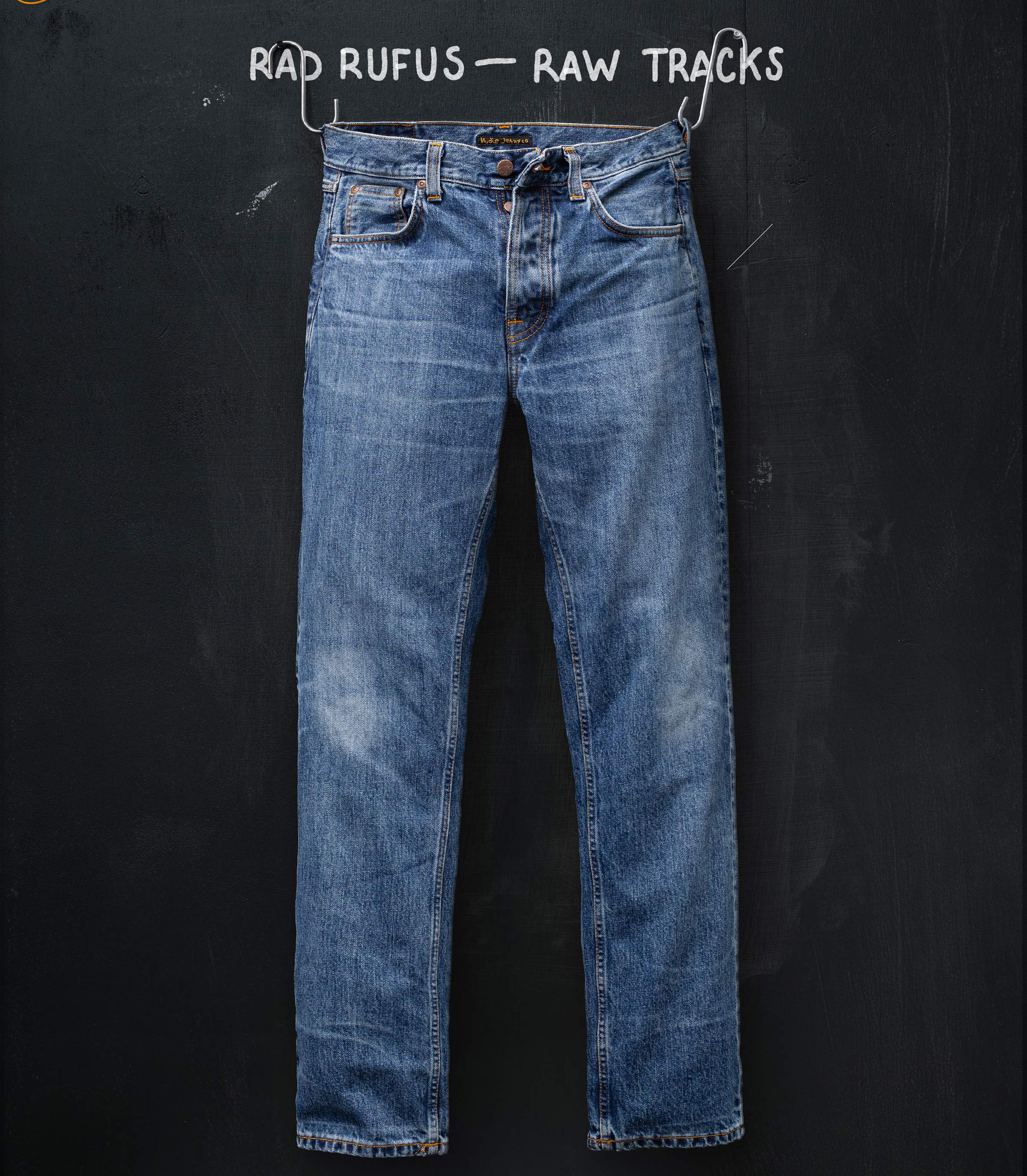 Nudie Jeans Rad Rufus Raw Tracks worn-in blue jeans on a wire hanger against a chalkboard background.