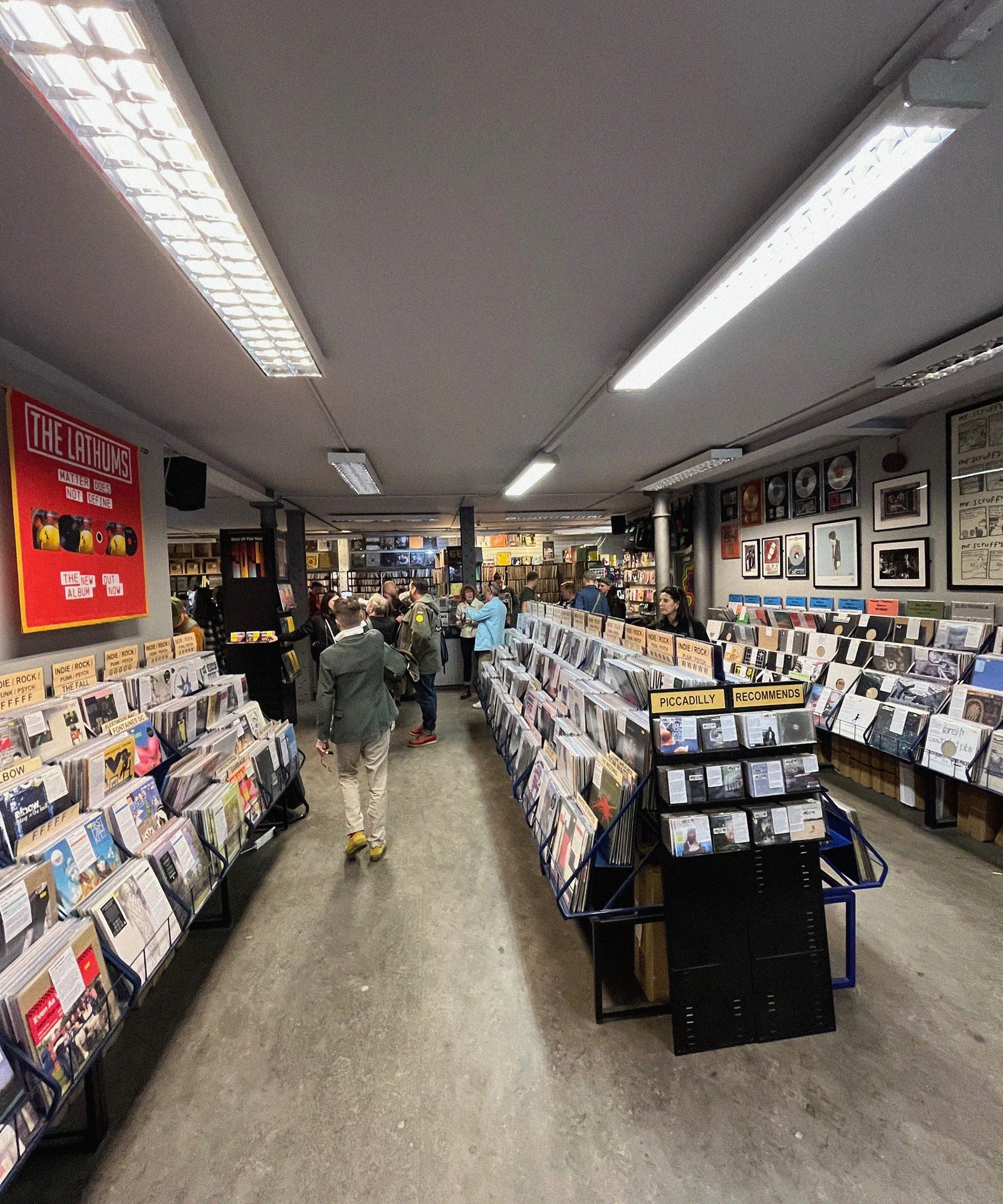 A bustling record store with rows of vinyl records, posters on the walls, and people browsing. Bright overhead lighting illuminates the space.