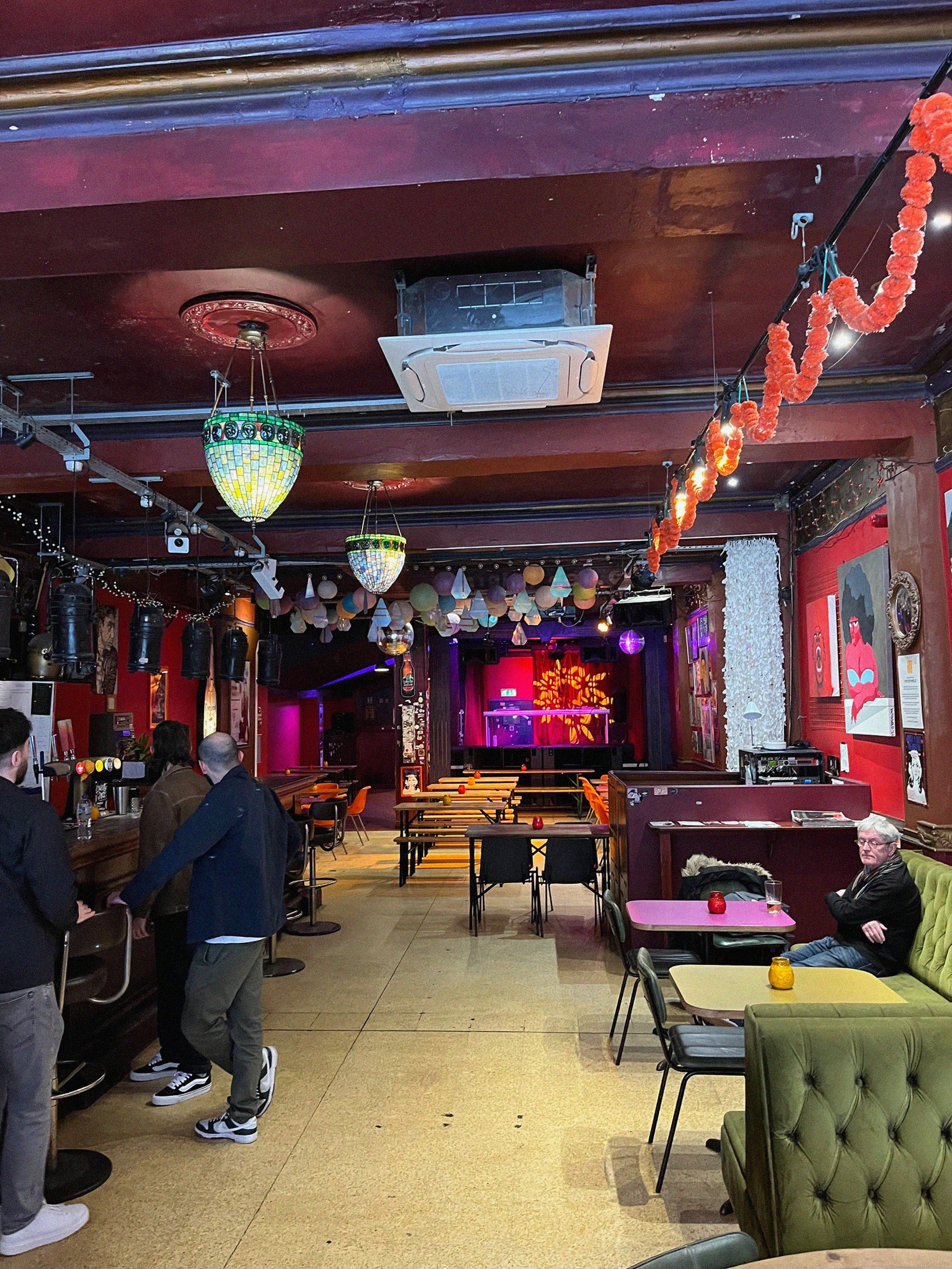A cozy bar with colorful lighting, chandeliers, and balloons. People are seated and standing, with a stage visible in the background.