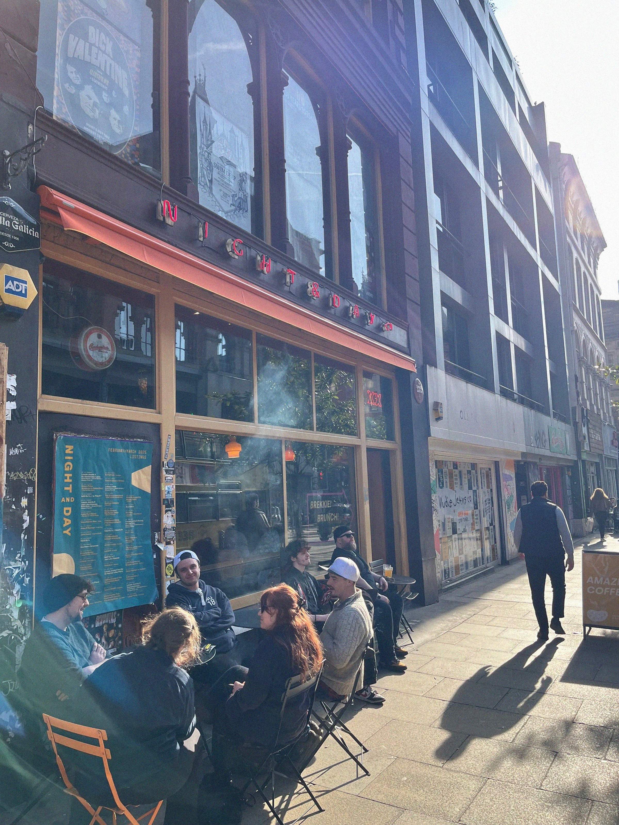 People sitting at outdoor tables of a café on a sunny street, with a menu displayed on the window and a person walking by.