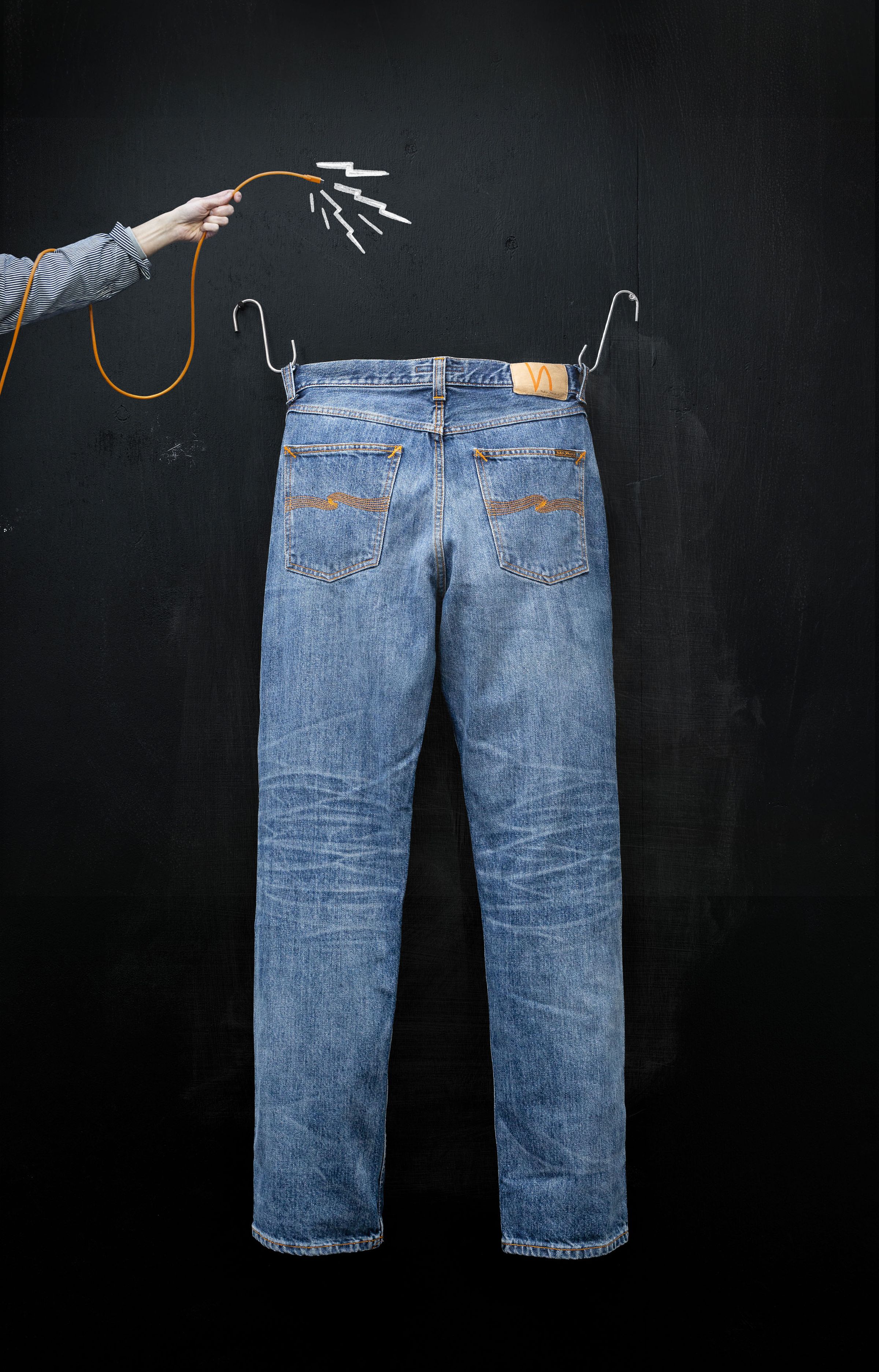 A pair of blue jeans hanging on a black background, with a hand holding an orange cable connected to the jeans' waistband.
