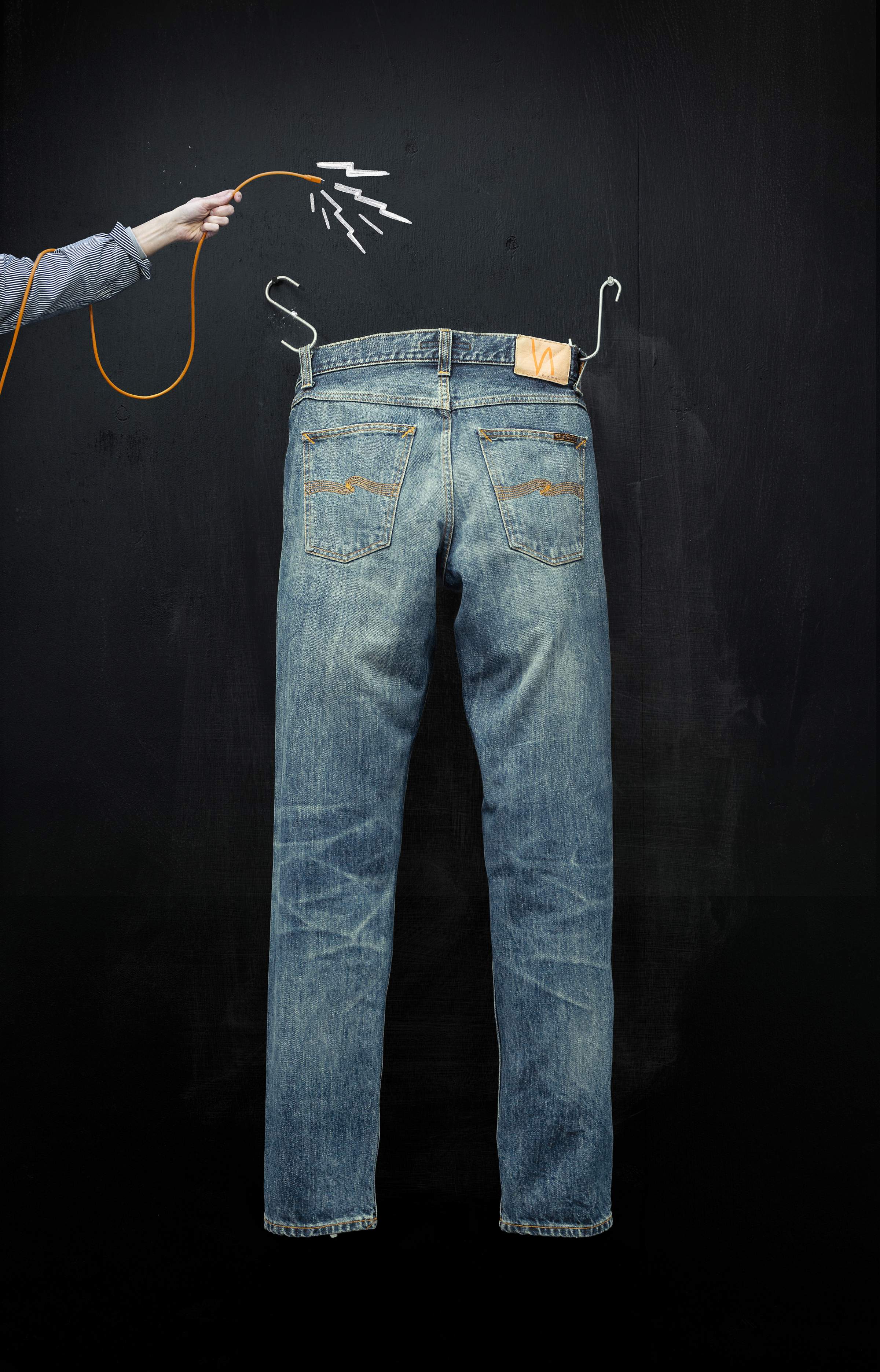 A pair of jeans hanging on a black wall, with a hand holding an orange cable connected to the jeans, resembling a charging setup.