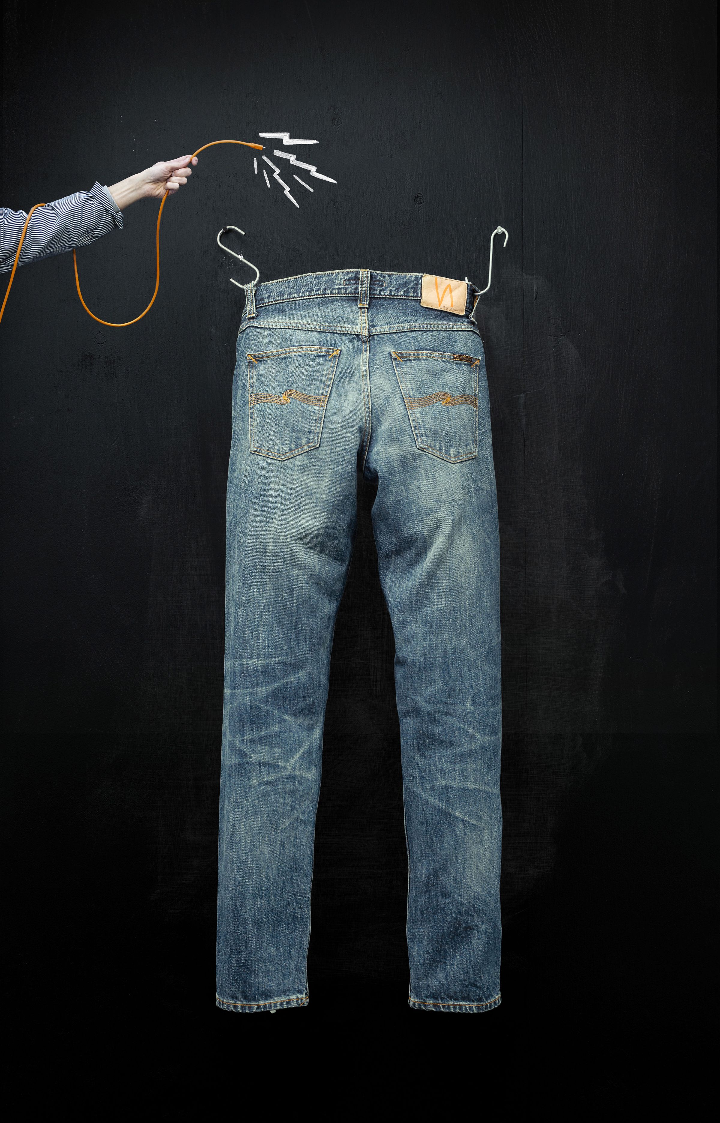 A pair of jeans hanging on a black wall, with a hand holding an orange cable connected to the jeans, resembling a charging setup.