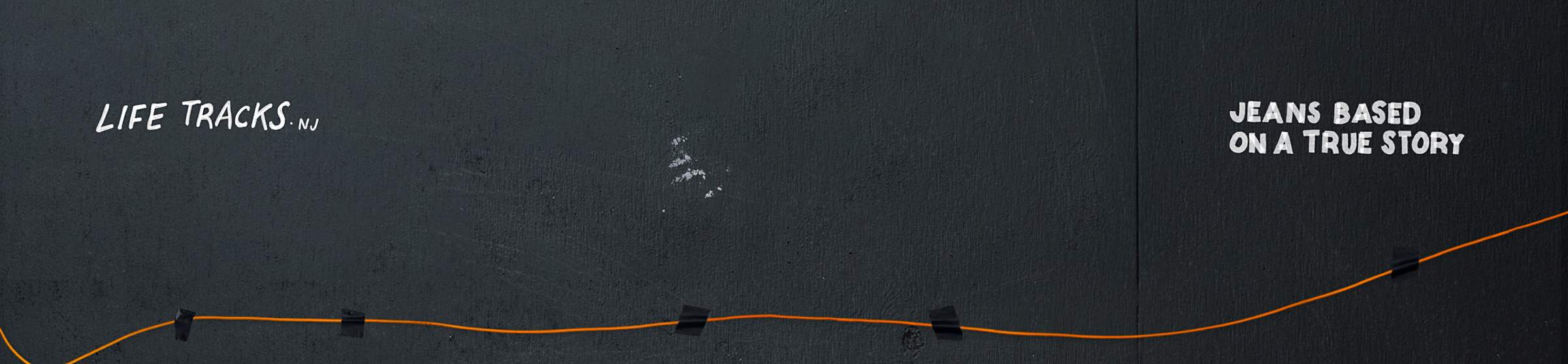 Black wall with an orange line taped across. Text: "Life Tracks NJ" on the left and "Jeans Based on a True Story" on the right.