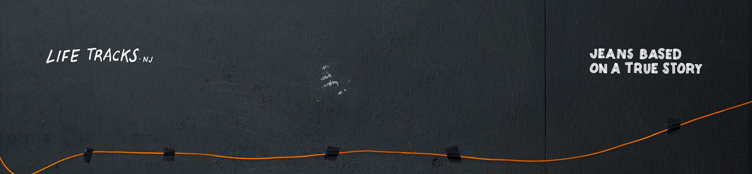 Black wall with an orange line taped across. Text: "Life Tracks NJ" on the left and "Jeans Based on a True Story" on the right.
