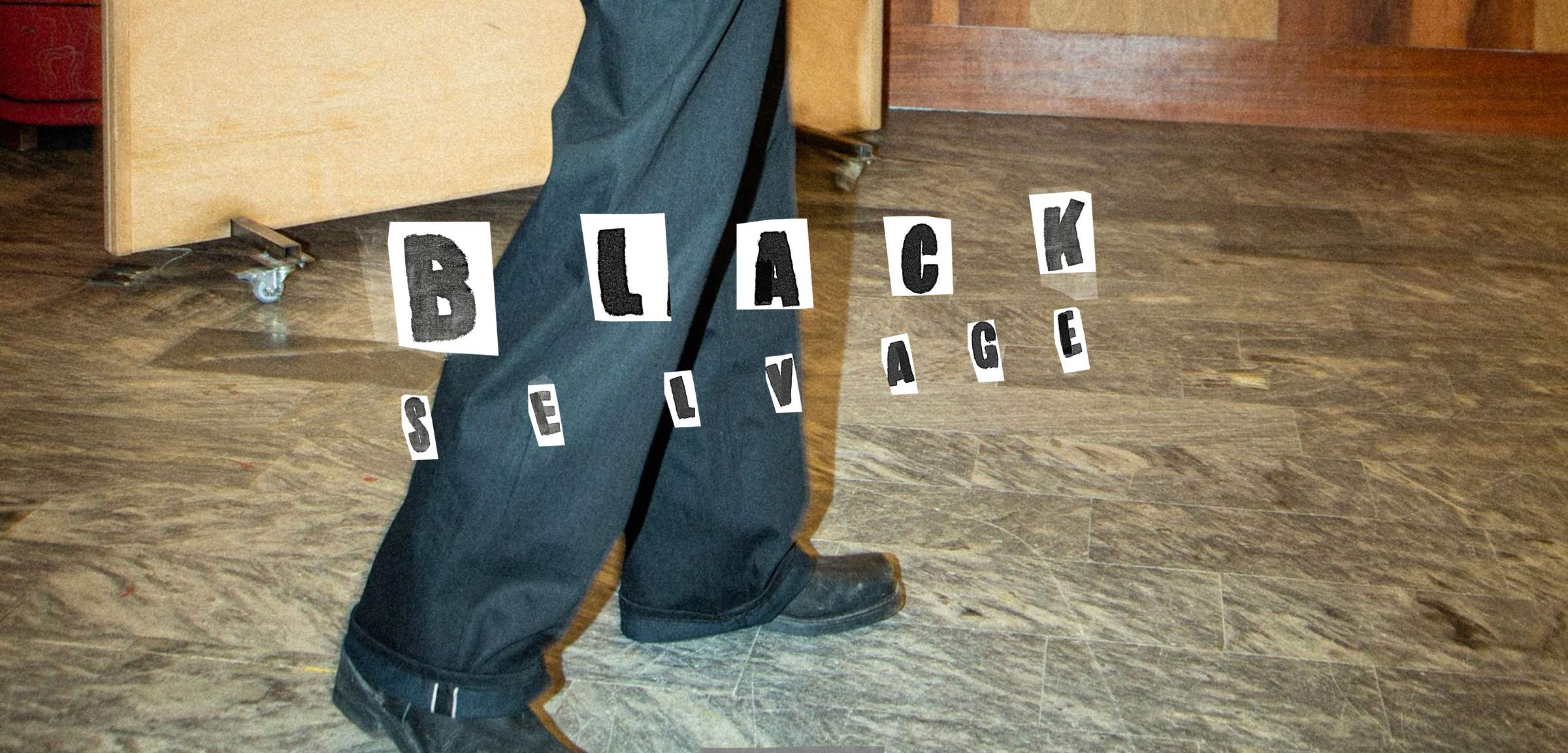 Model in Nudie Jeans Black Selvedge jeans with rolled cuffs and black shoes, "Black Selvedge" cut-out letter graphic.