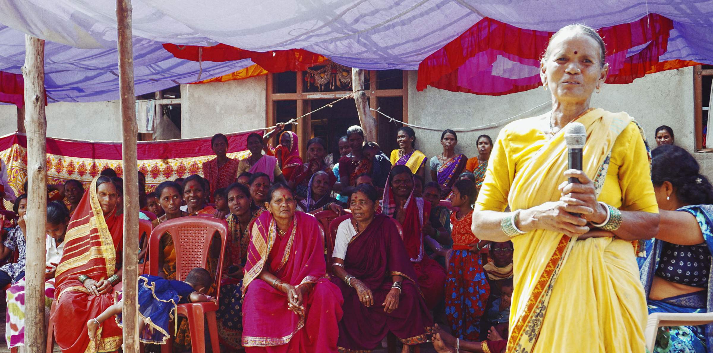 An elderly woman in a yellow sari speaks into a microphone, surrounded by seated women under a colorful canopy.