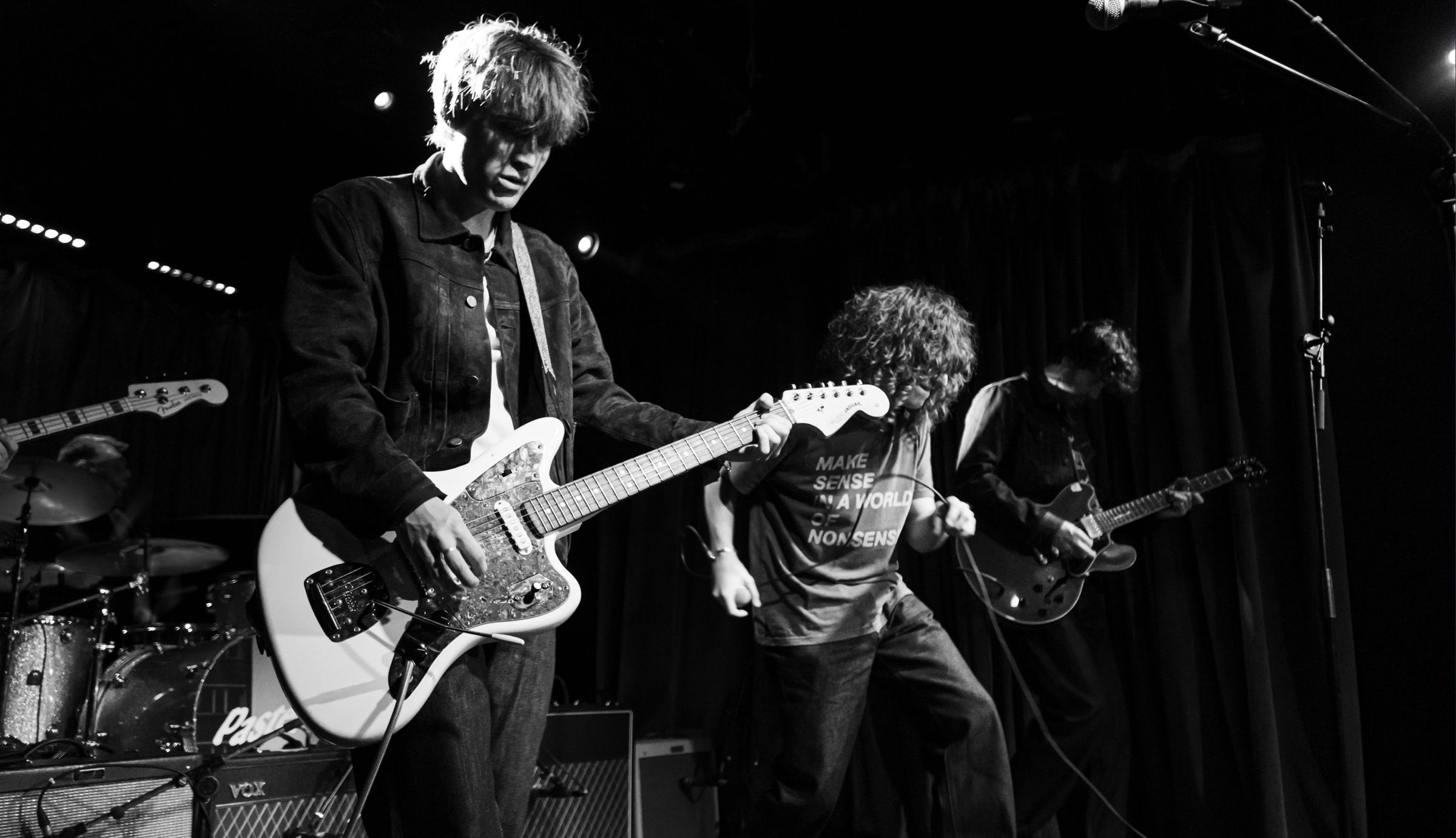 Black-and-white photo of a Pastel on stage — guitarist with white electric guitar, curly-haired vocalist mid-move, second guitarist and drummer behind.