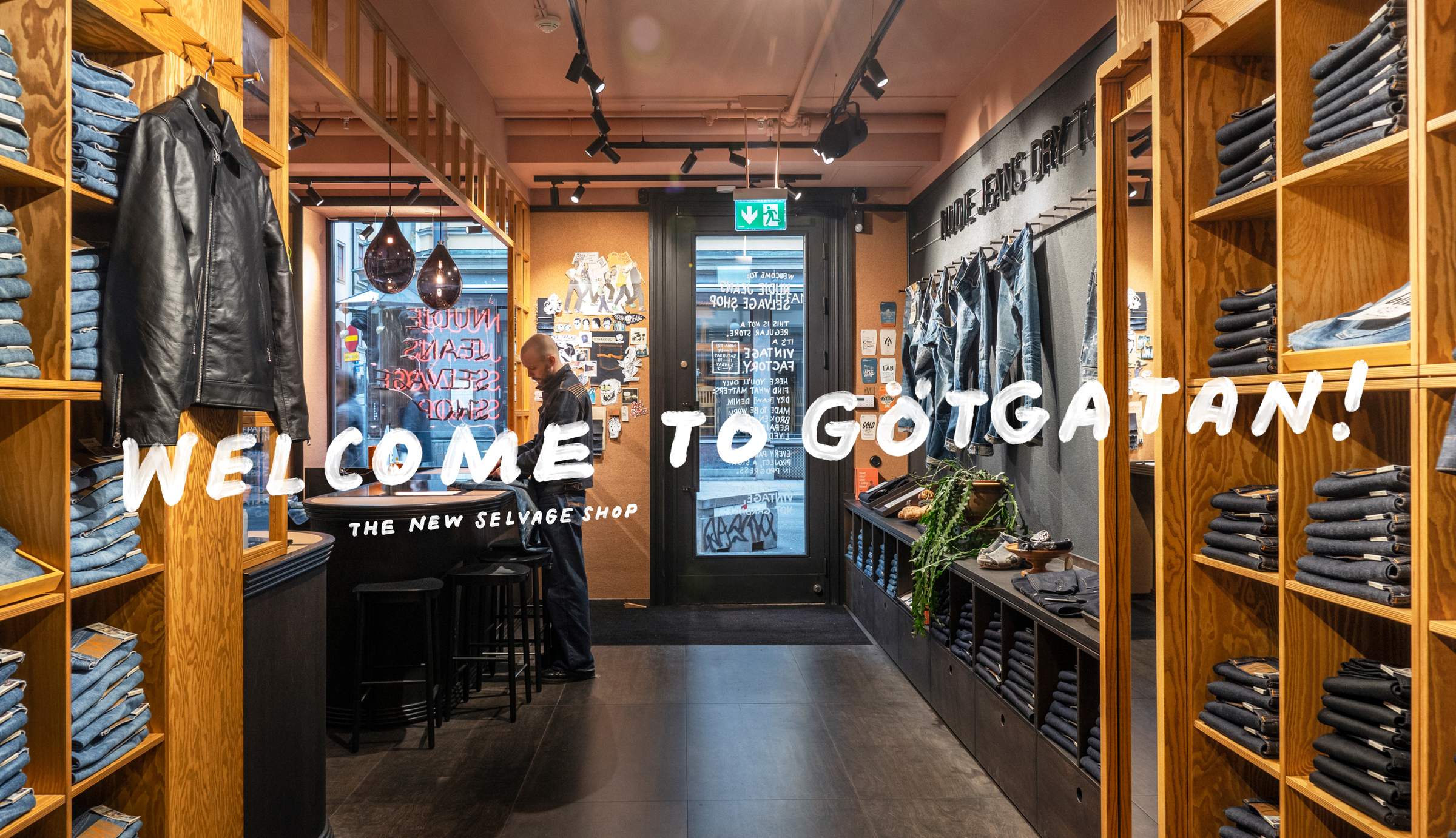 Clothing store interior with wooden shelves displaying jeans and jackets. "WELCOME TO GÖTGATAN! THE NEW SELVAGE SHOP" text overlay.