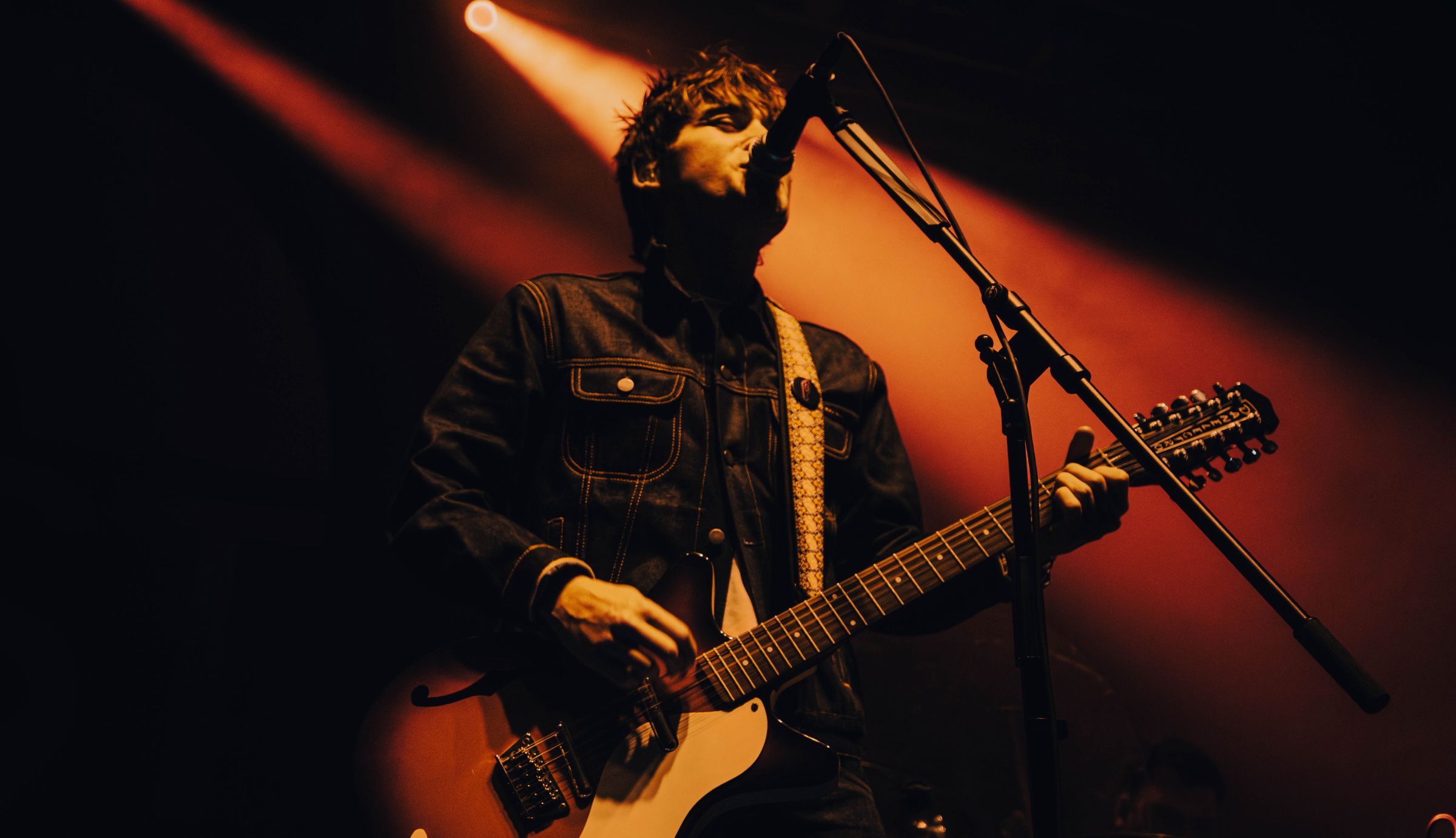 Musician in denim jacket playing electric guitar and singing into a microphone on stage, warm orange spotlight behind.