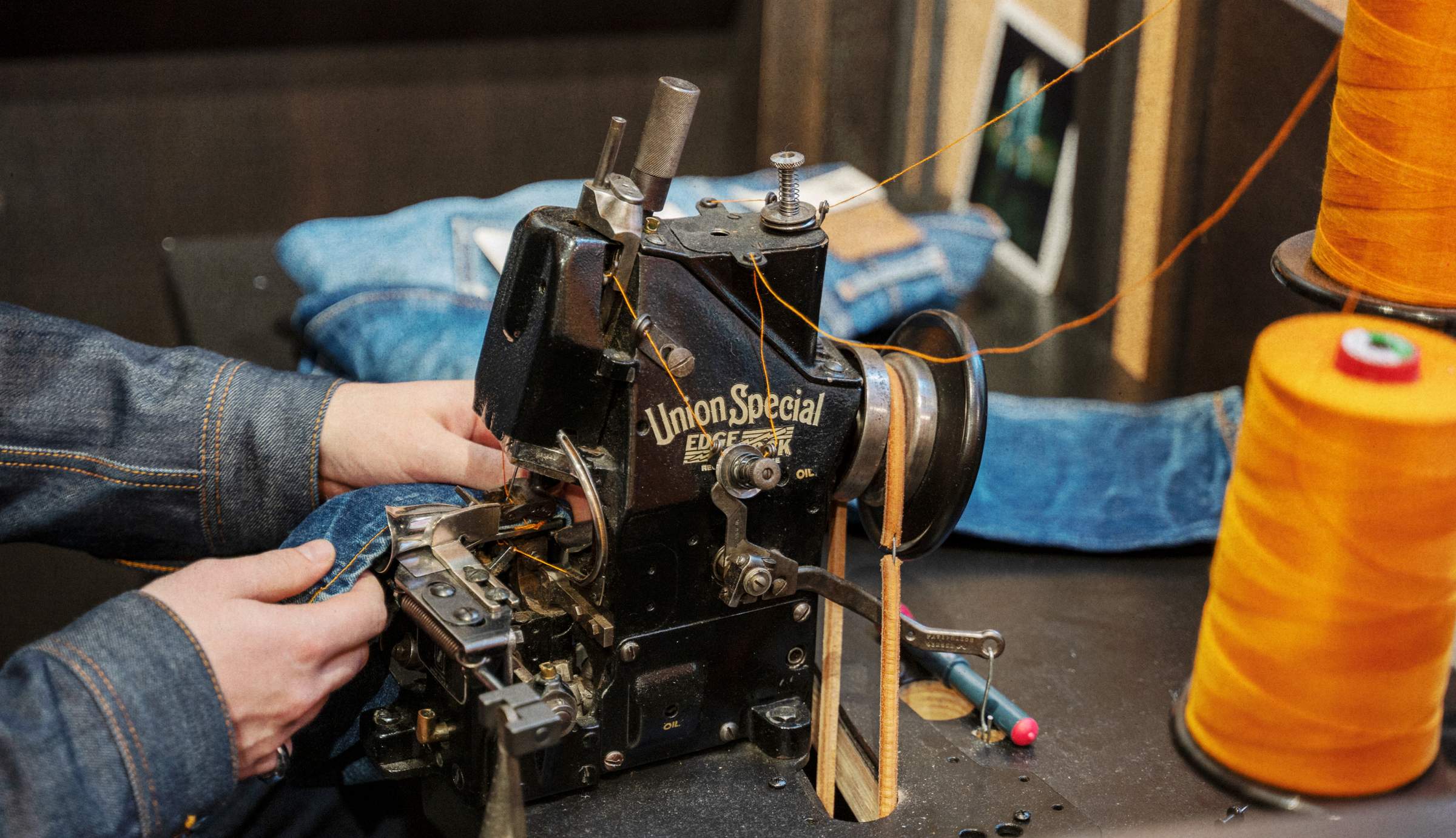Close-up of hands sewing blue denim fabric on a vintage Union Special sewing machine surrounded by spools of orange thread.