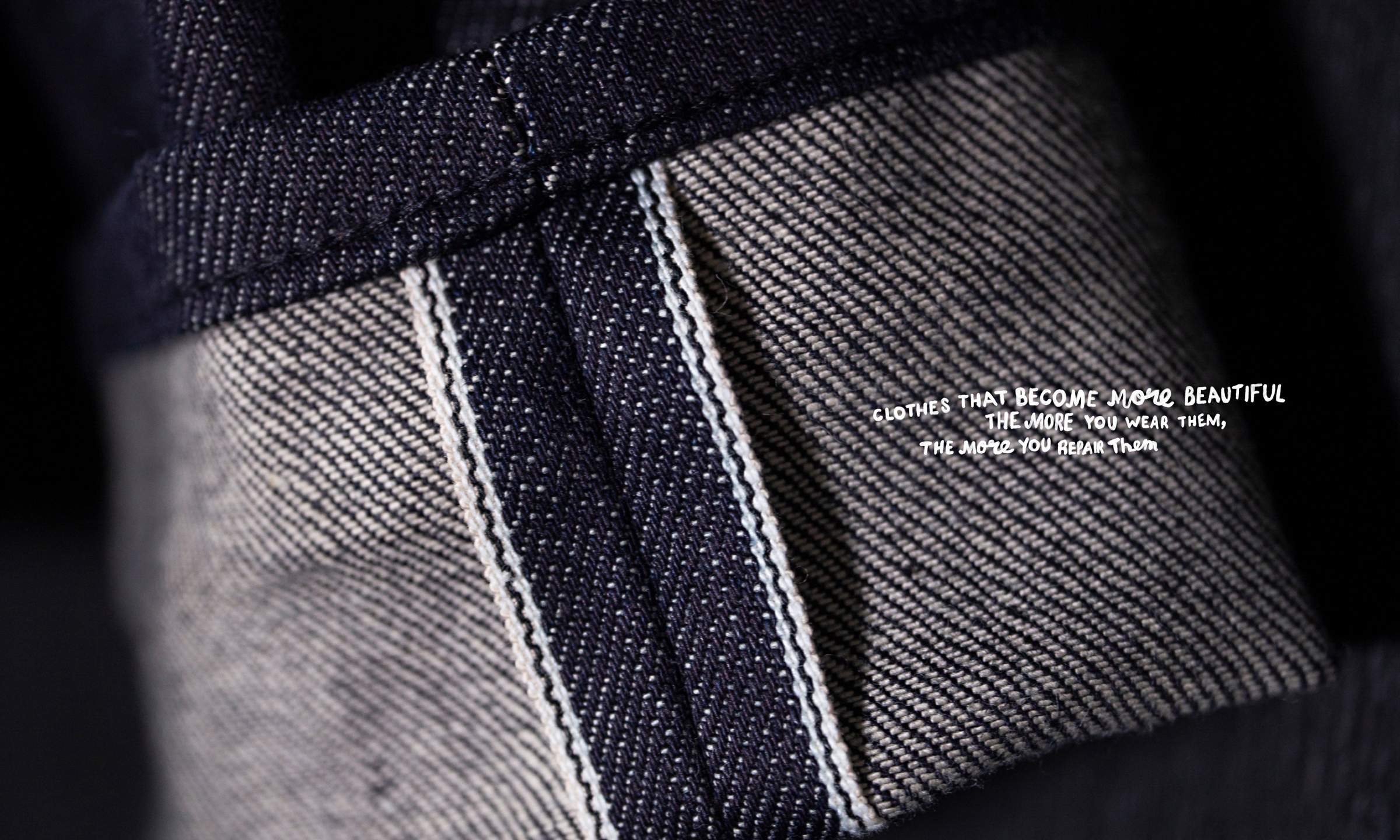 Close-up of dark denim fabric with visible stitching and a small white text: "Clothes that become more beautiful the more you wear them, the more you repair them."