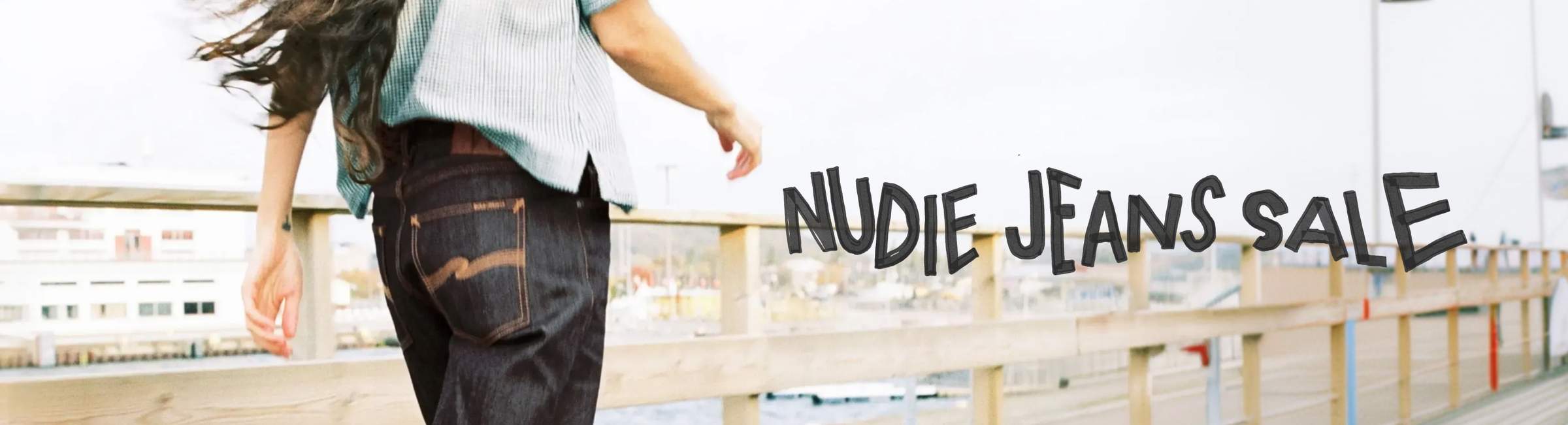 Person in denim and shirt walking on a wooden bridge with urban background, "Nudie Jeans Sale" text overlay.
