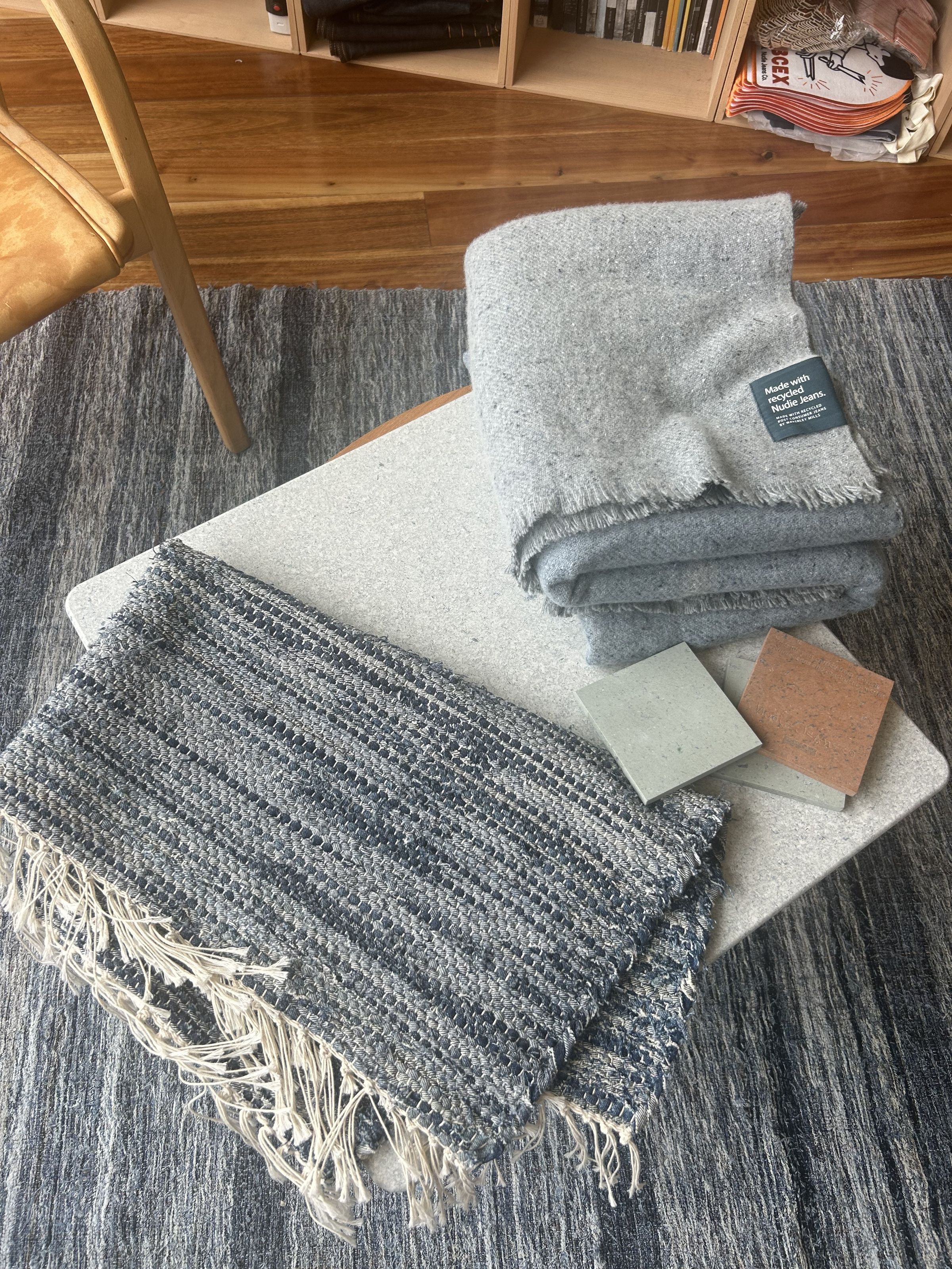 A small table with folded gray blankets, a woven rug, and two square tiles, set on a gray carpeted floor near a wooden chair and bookshelf.