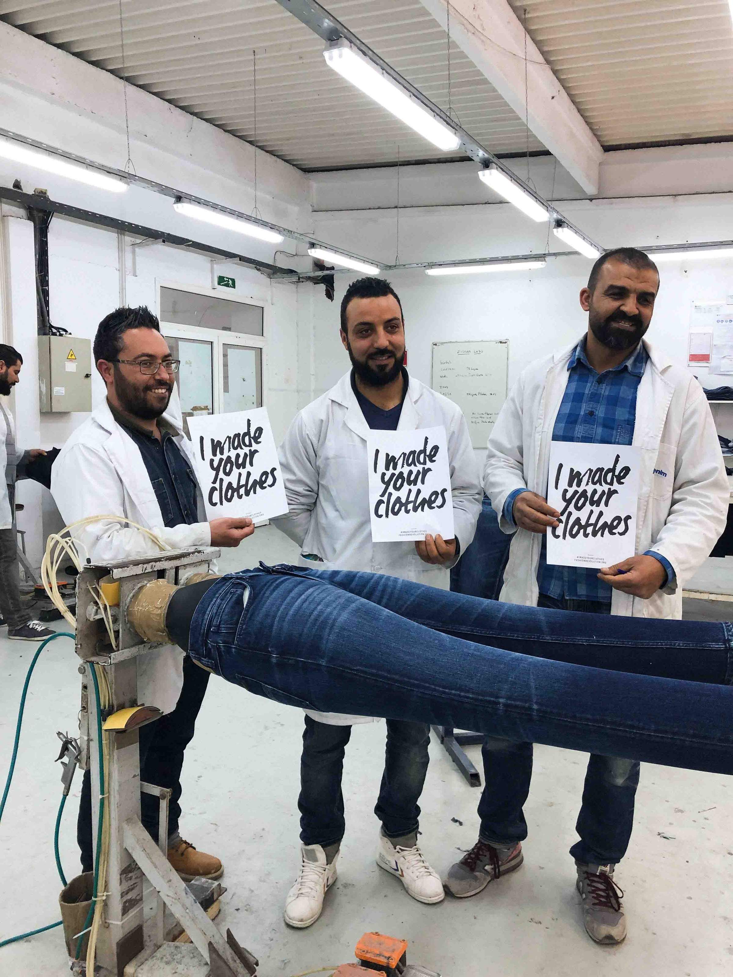 Three men in a workshop hold signs saying "I made your clothes" next to a machine with denim jeans, highlighting garment production.