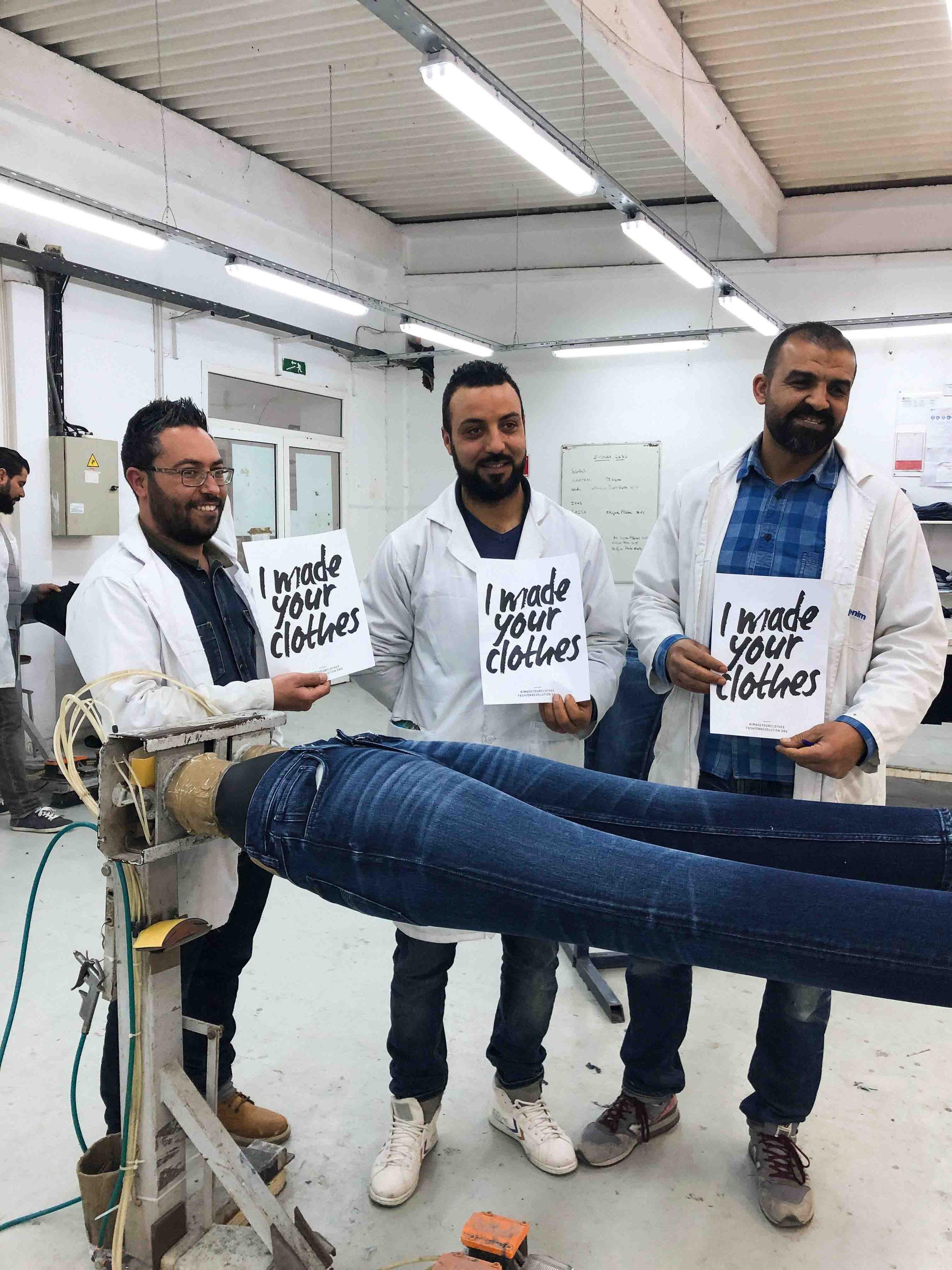 Three men in a workshop hold signs saying "I made your clothes" next to a machine with denim jeans, highlighting garment production.