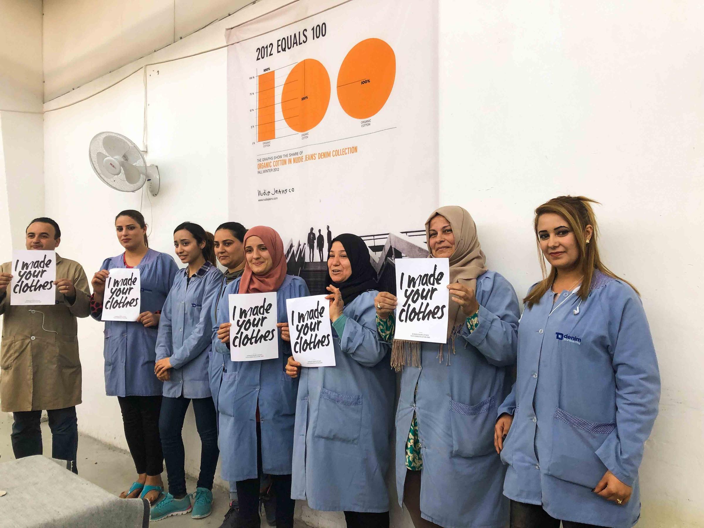 A group of people in blue coats hold signs reading "I made your clothes" in front of a wall with a chart.