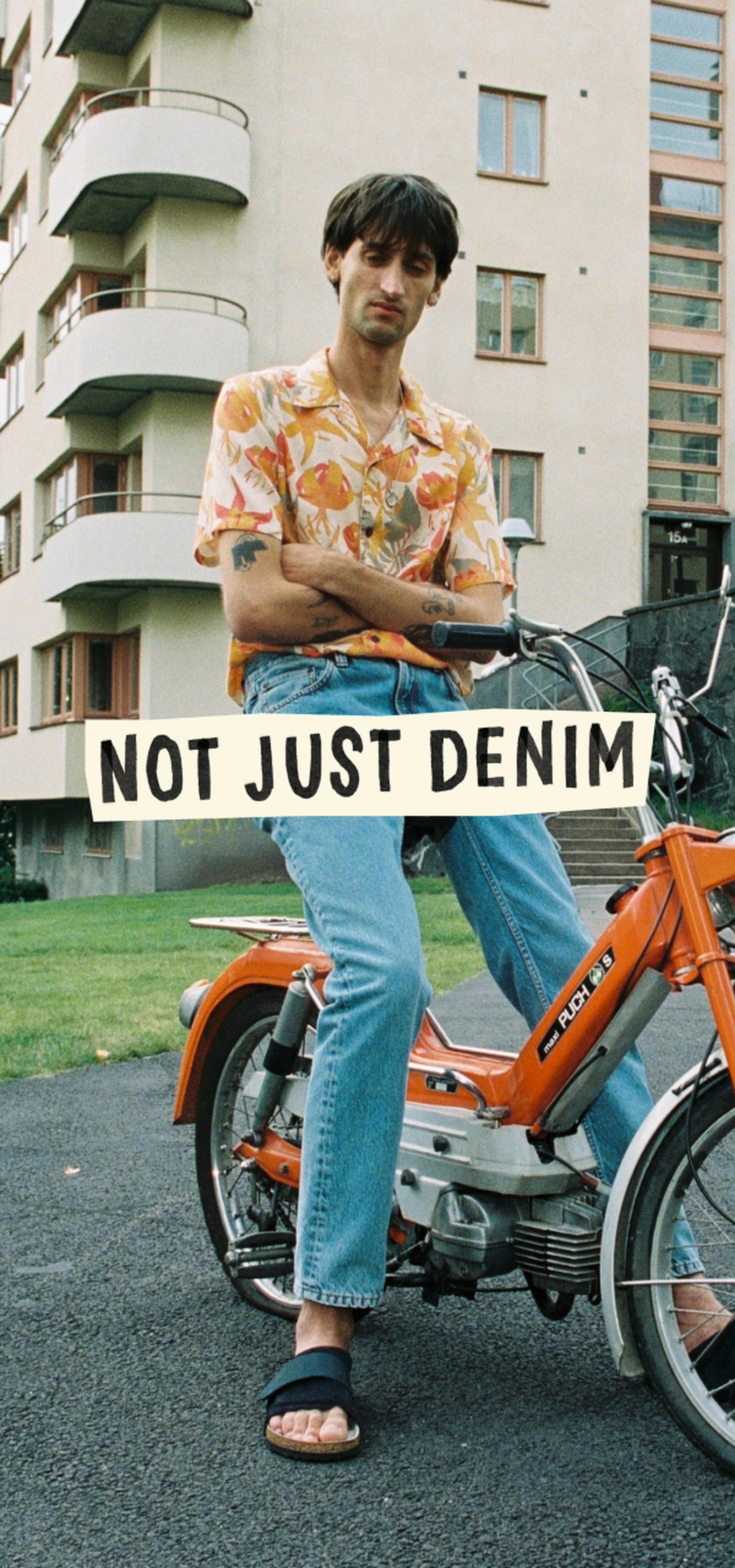 Man in a floral shirt and jeans sits on an orange moped in front of an apartment building, with a "Not Just Denim" sign overlay.