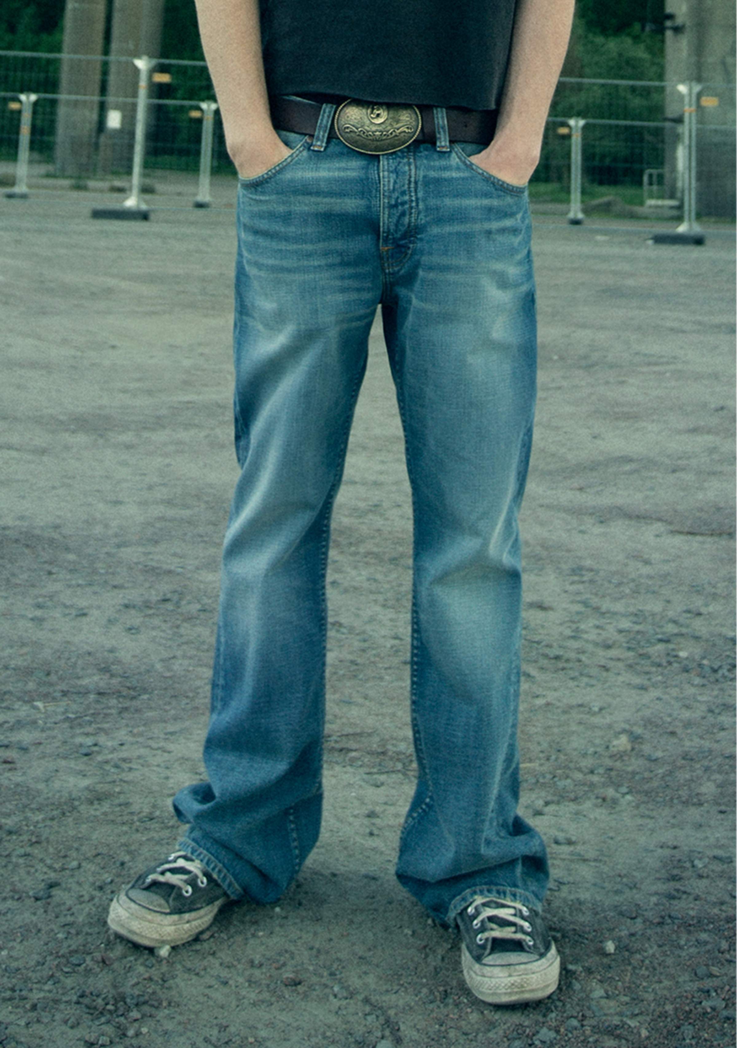 Hannes in a pair of Flare Glenn and sneakers stands on gravel, hands in pockets, wearing a belt with a large buckle.