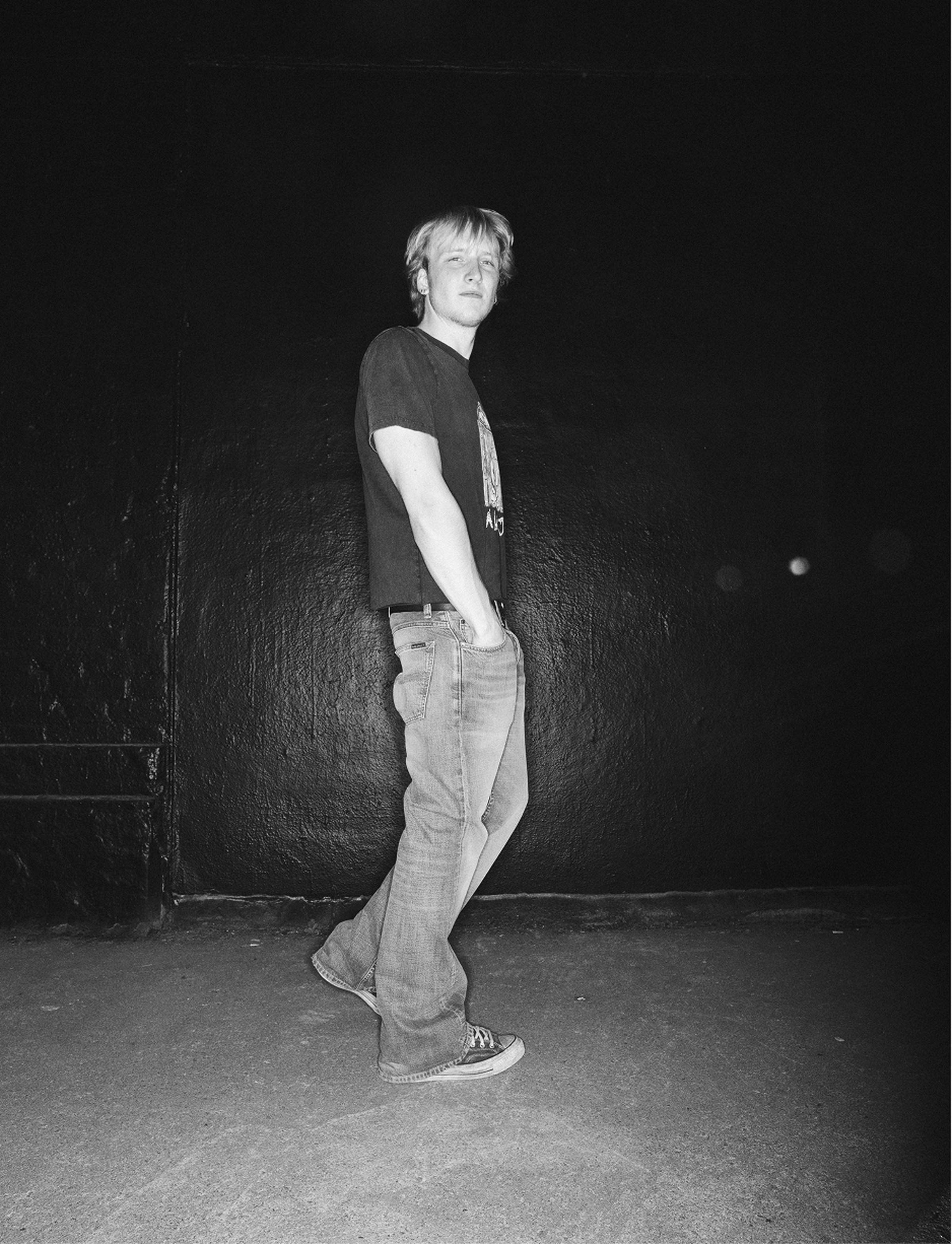 Hannes in a pair of Flare Glenn and sneakers stands on gravel, hands in pockets, wearing a belt with a large buckle.