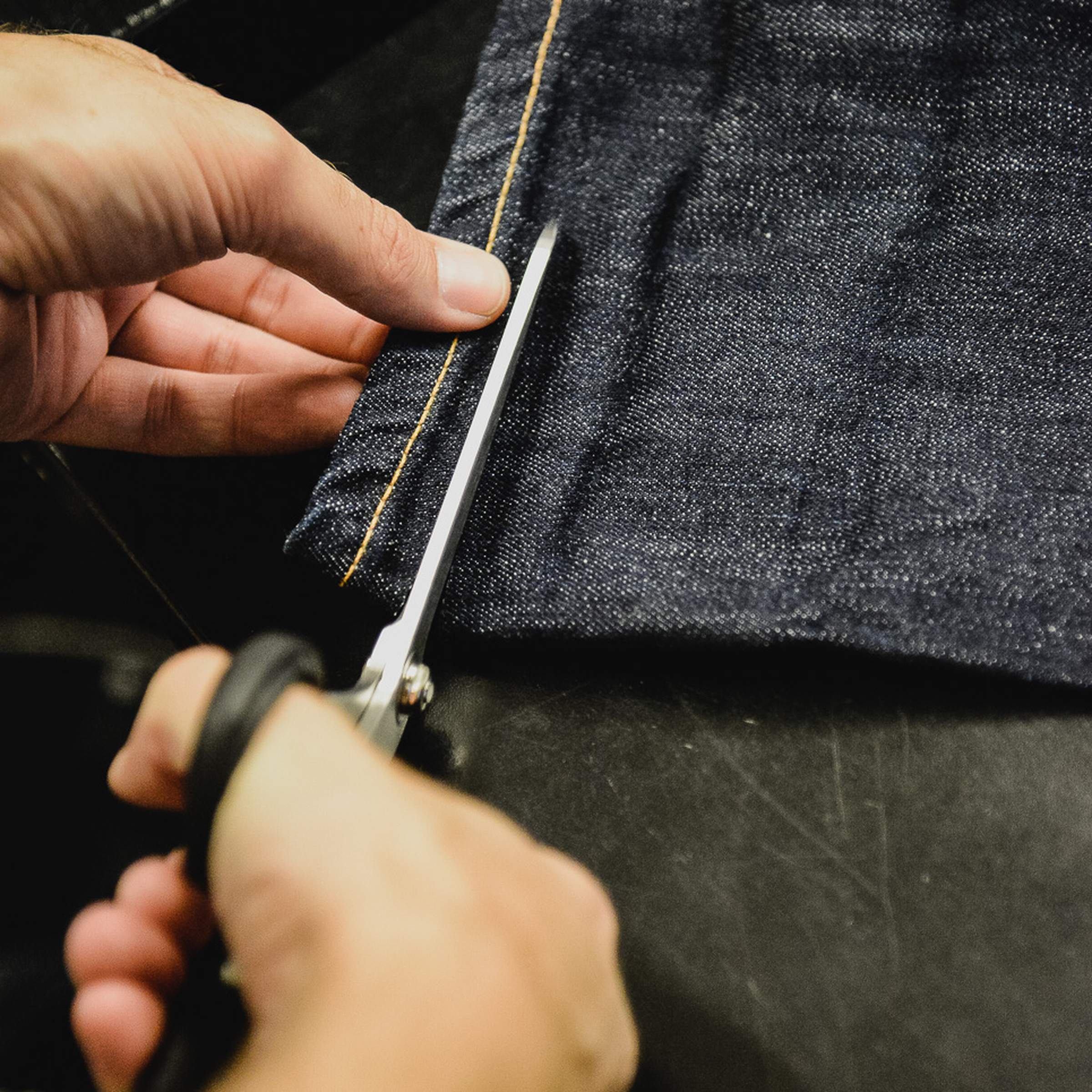 Hands cutting dark denim fabric with scissors on a black surface.