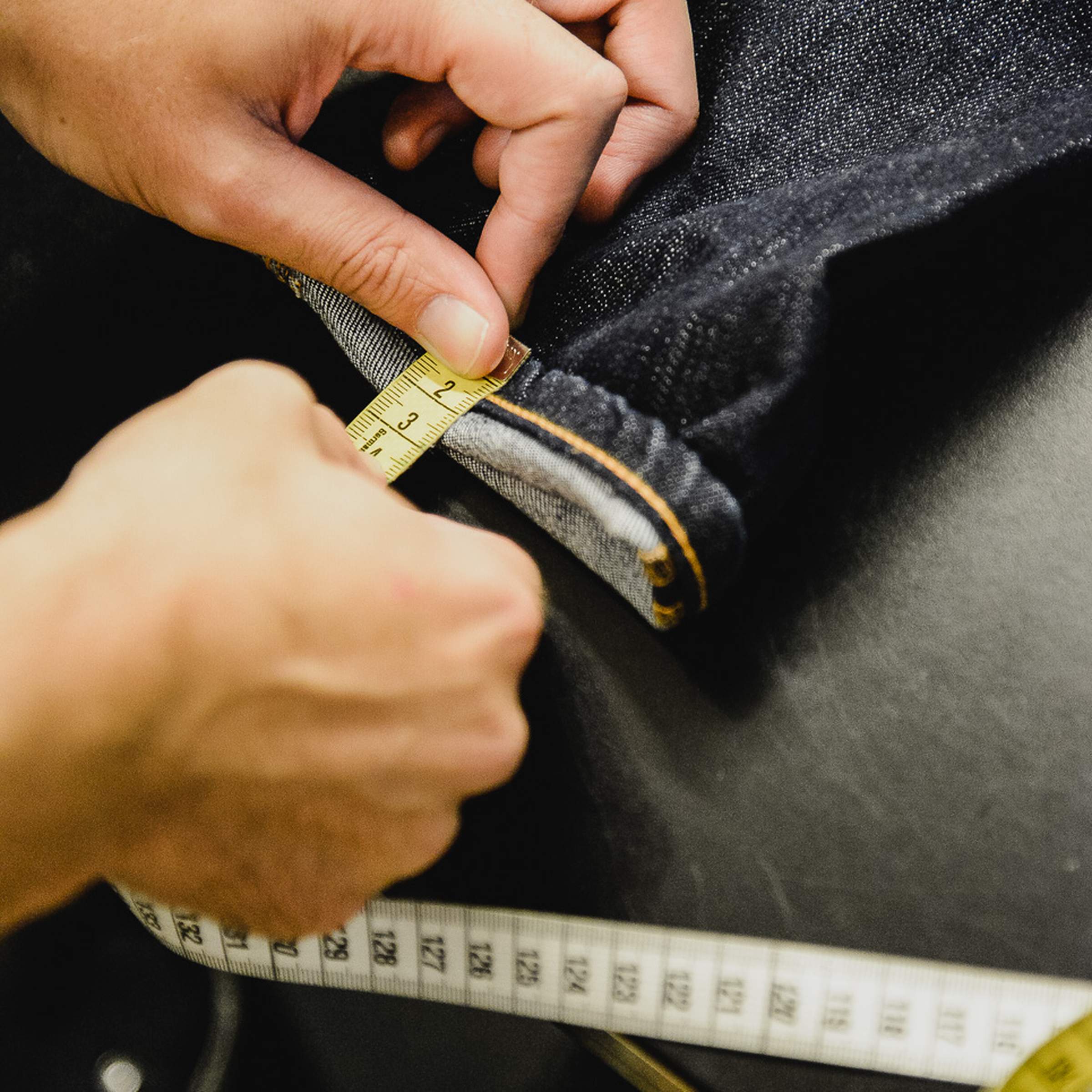 Hands measuring denim fabric with a tape measure on a table, focusing on precise tailoring.