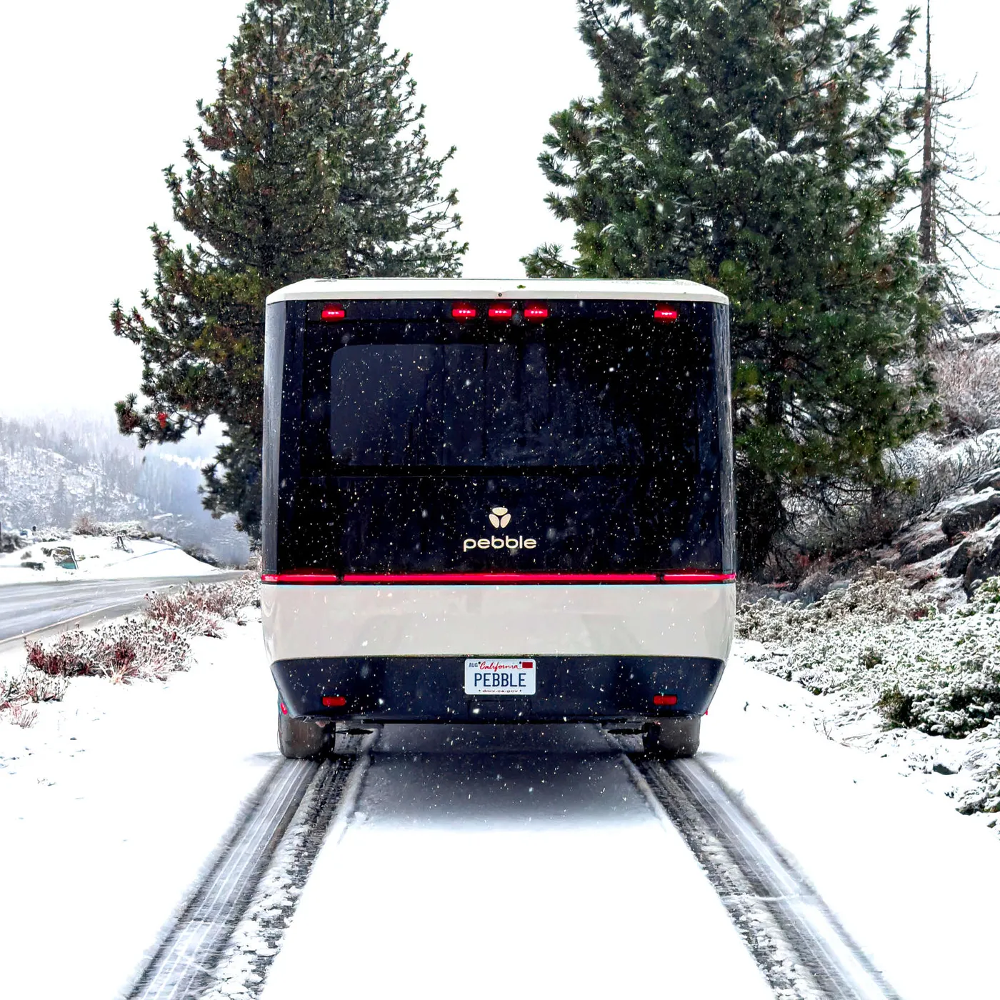A Pebble Flow on a snowy road surrounded by pine trees and a snowy landscape, leaving tire tracks in the fresh snow.