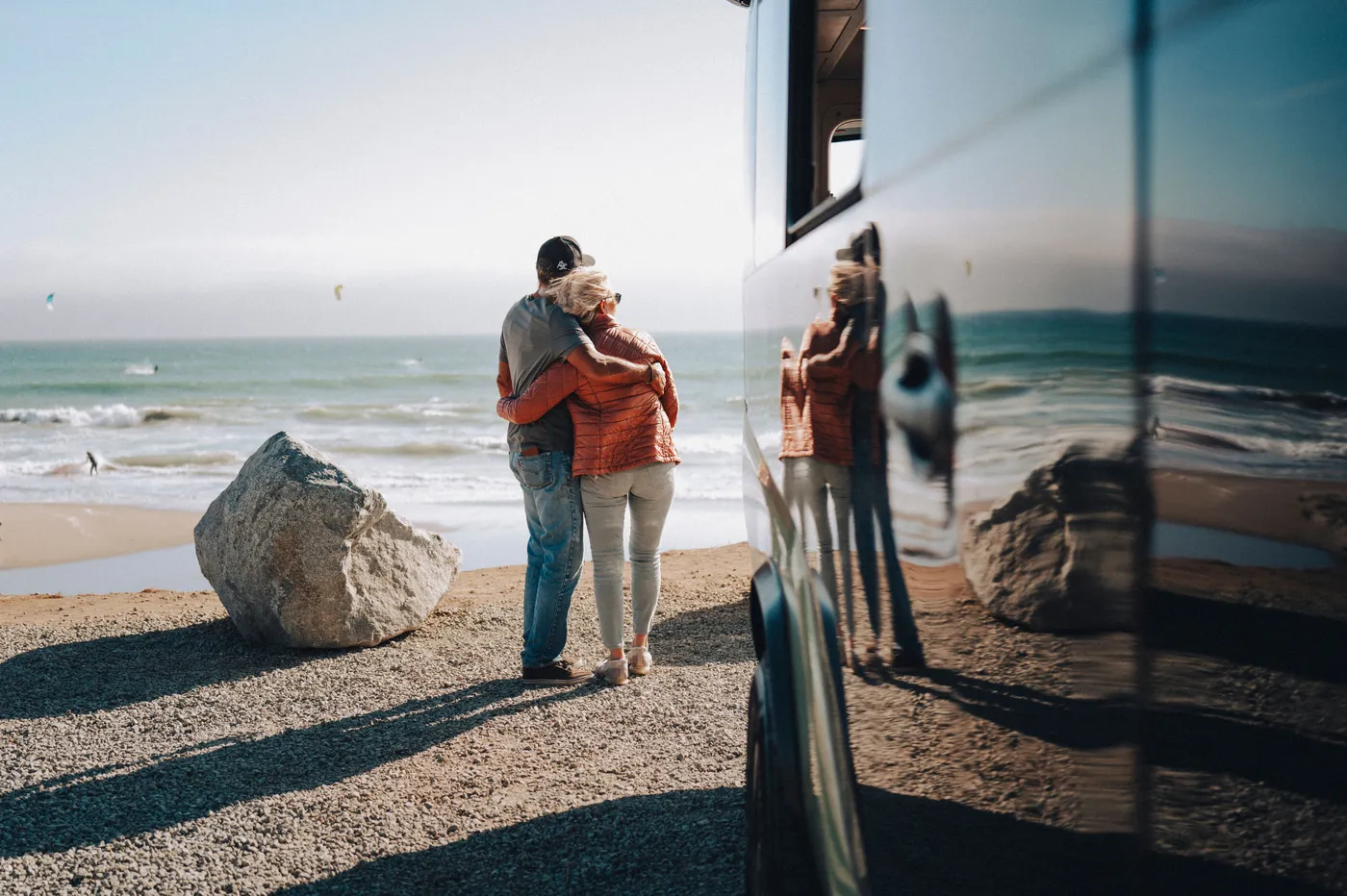 A couple embraces on a beach, facing the ocean. Their reflection is visible on the Pebble Flow's shiny surface, with a large rock in the foreground.