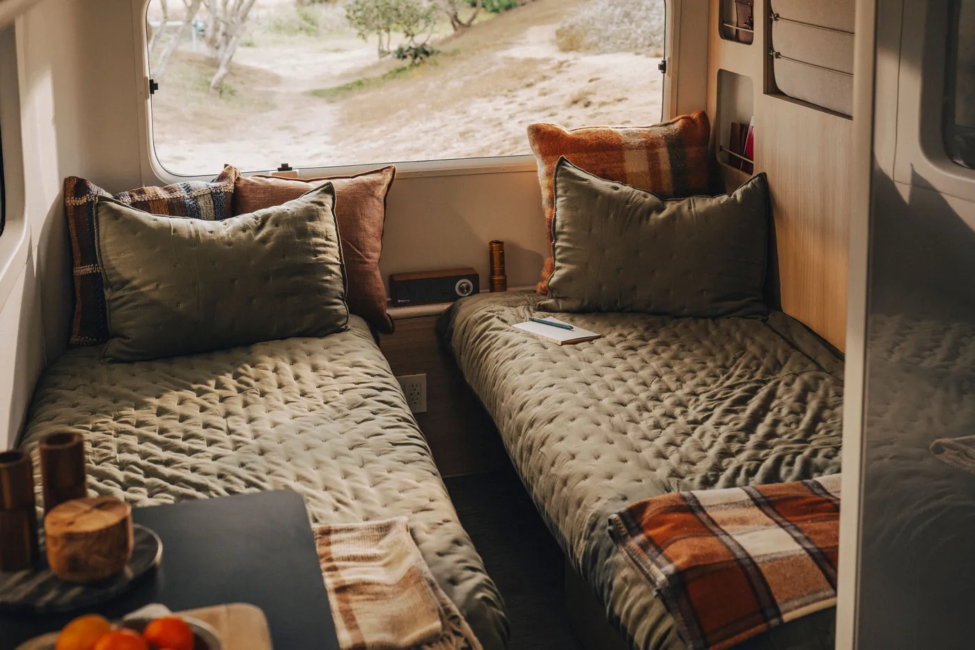 Cozy interior of a Pebble Flow with two twin beds, olive green bedding, plaid pillows, and a view outside the window of a natural landscape.