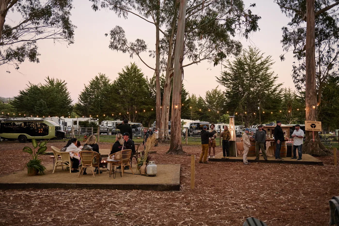Outdoor gathering with people sitting and socializing at tables under string lights among trees, beside parked Pebble Flows at dusk.