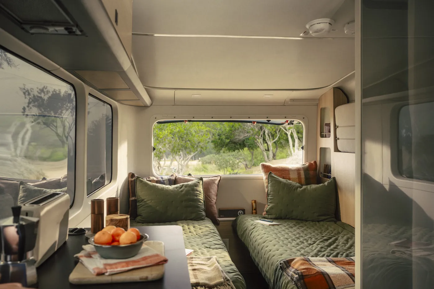 Cozy interior of a Pebble Flow with two twin beds, olive green bedding, plaid pillows, and a view outside the window of a natural landscape.