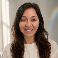 A woman with long brown hair smiles warmly, standing in soft natural light with a slight rainbow visible in the background.